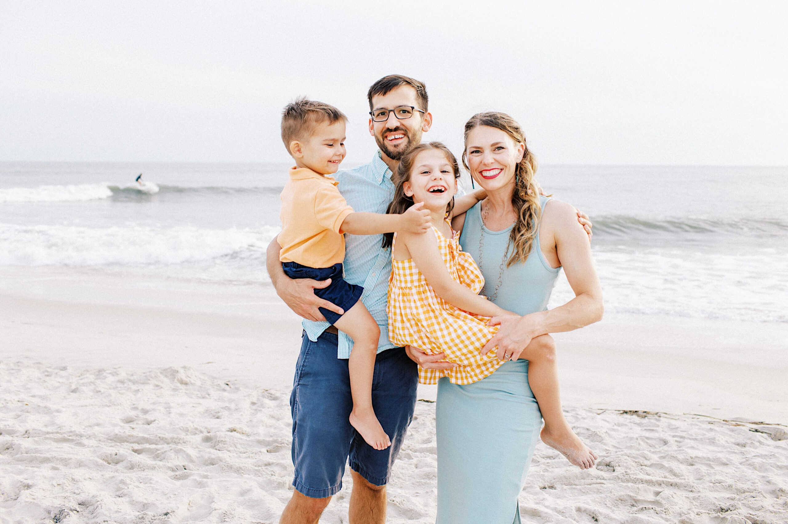 A family of four stands on a sandy beach with the ocean in the background, smiling at the camera. The parents hold their two young children, capturing special family photos on Cape May Beach in New Jersey.