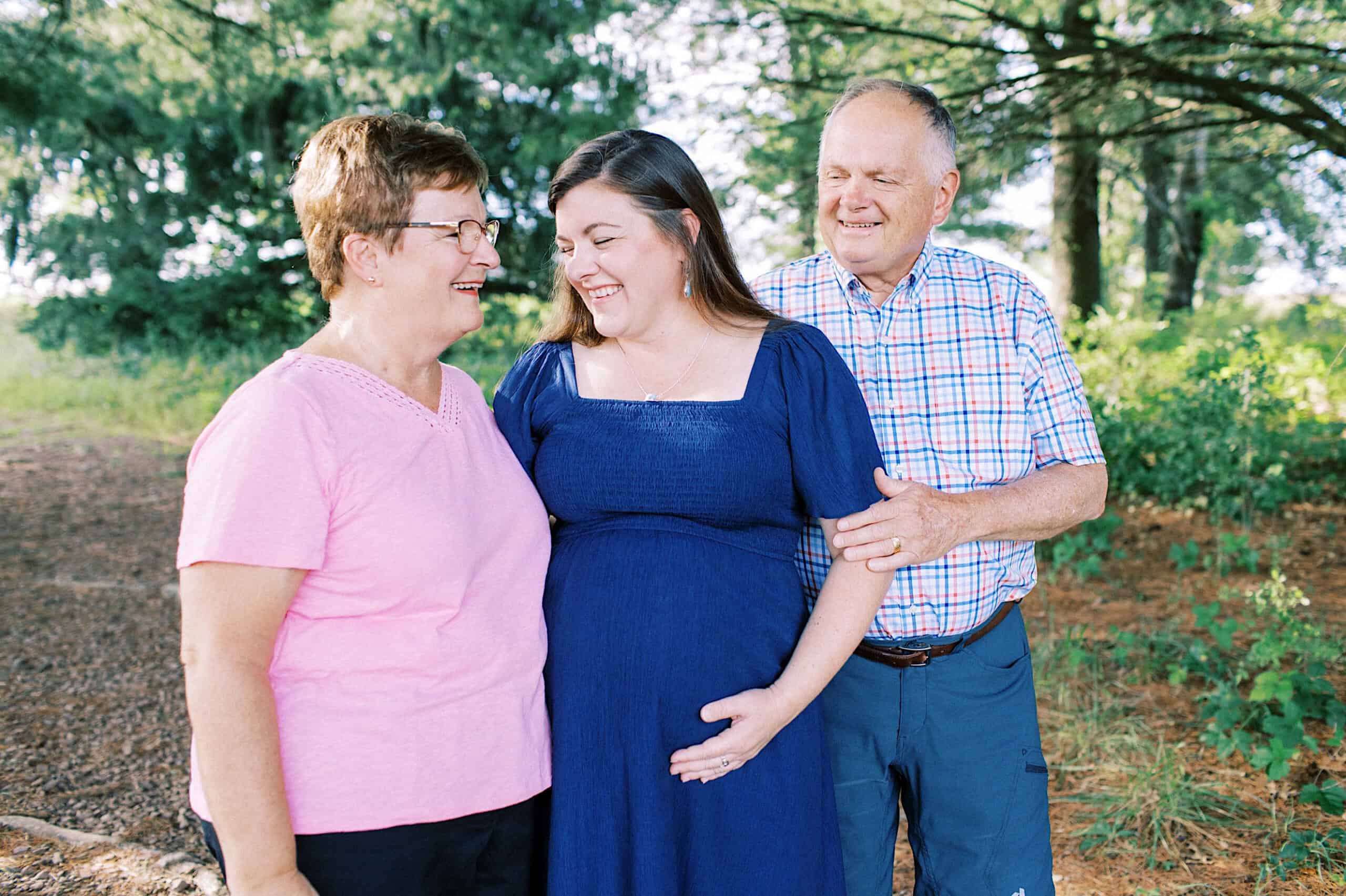 Three adults stand outdoors smiling for extended family photos; a pregnant woman in a blue dress is in the center, with an older woman in a pink shirt and an older man in a plaid shirt on either side, capturing a joyful maternity moment.