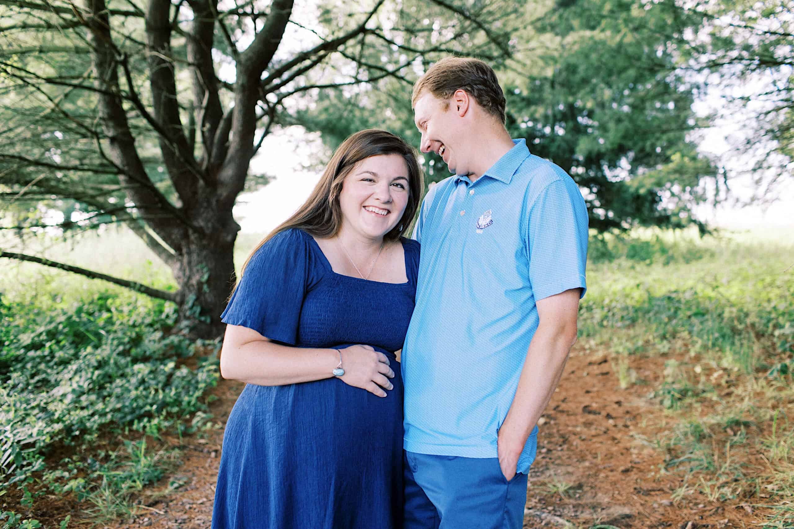 A woman in a blue dress stands smiling with her hand on her belly next to a man in a light blue shirt, outdoors in a wooded area—perfect for maternity or extended family photos.