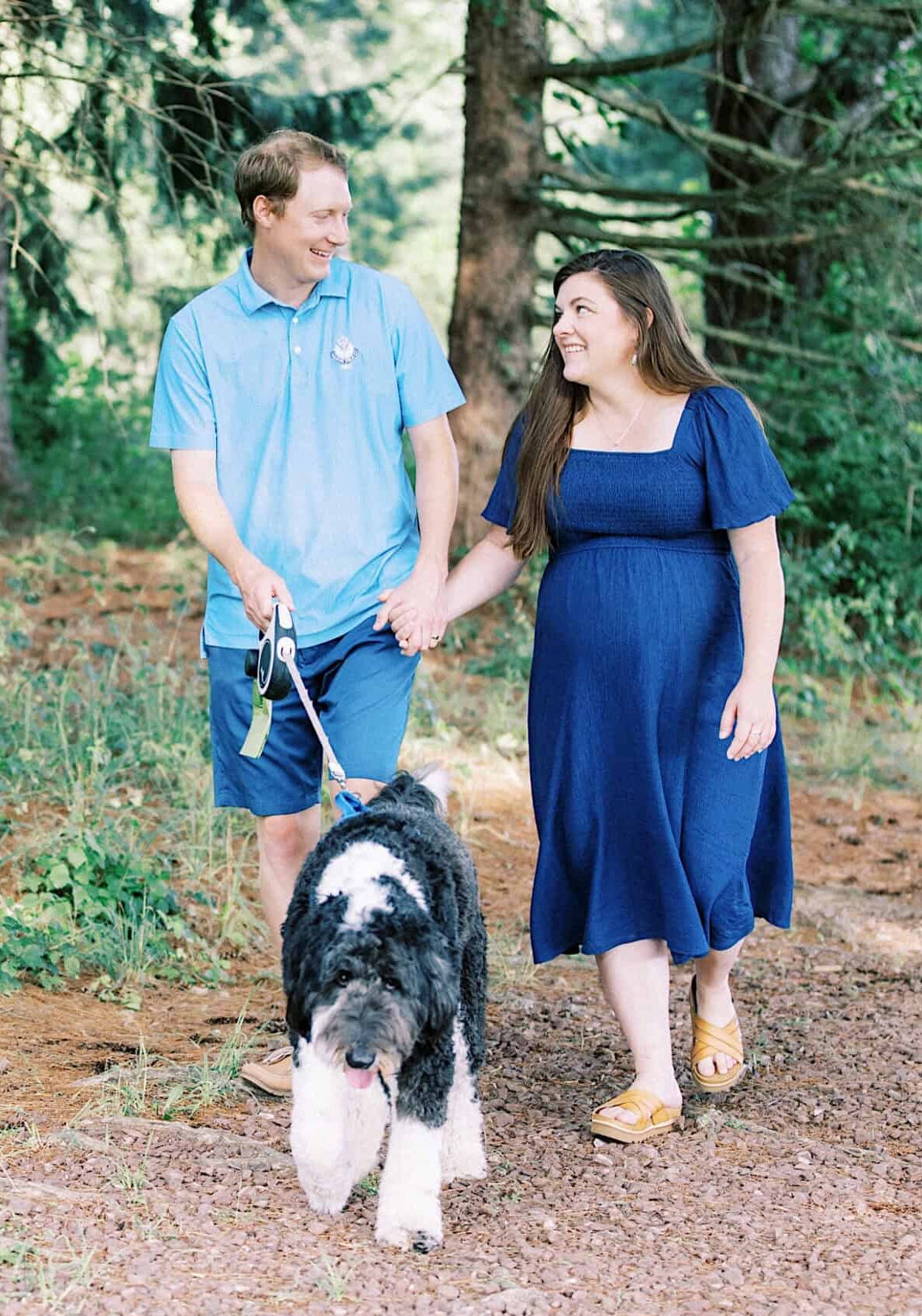 A man and a woman, smiling and holding hands, walk a large black and white dog on a dirt path through a wooded area—perfect for candid maternity and extended family photos.