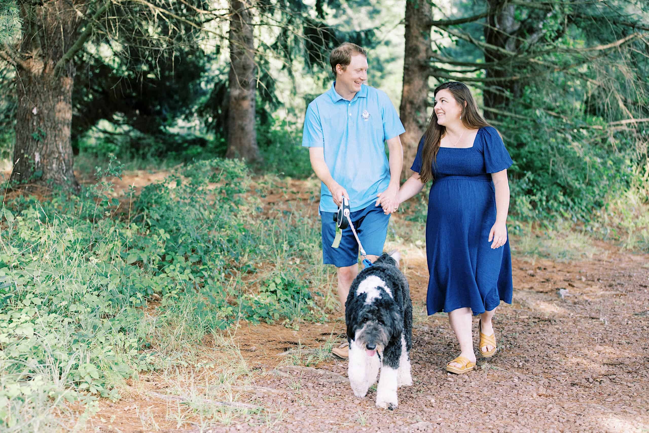 A man and a woman, smiling and holding hands, walk a large black and white dog on a dirt path through a wooded area—perfect for candid maternity and extended family photos.