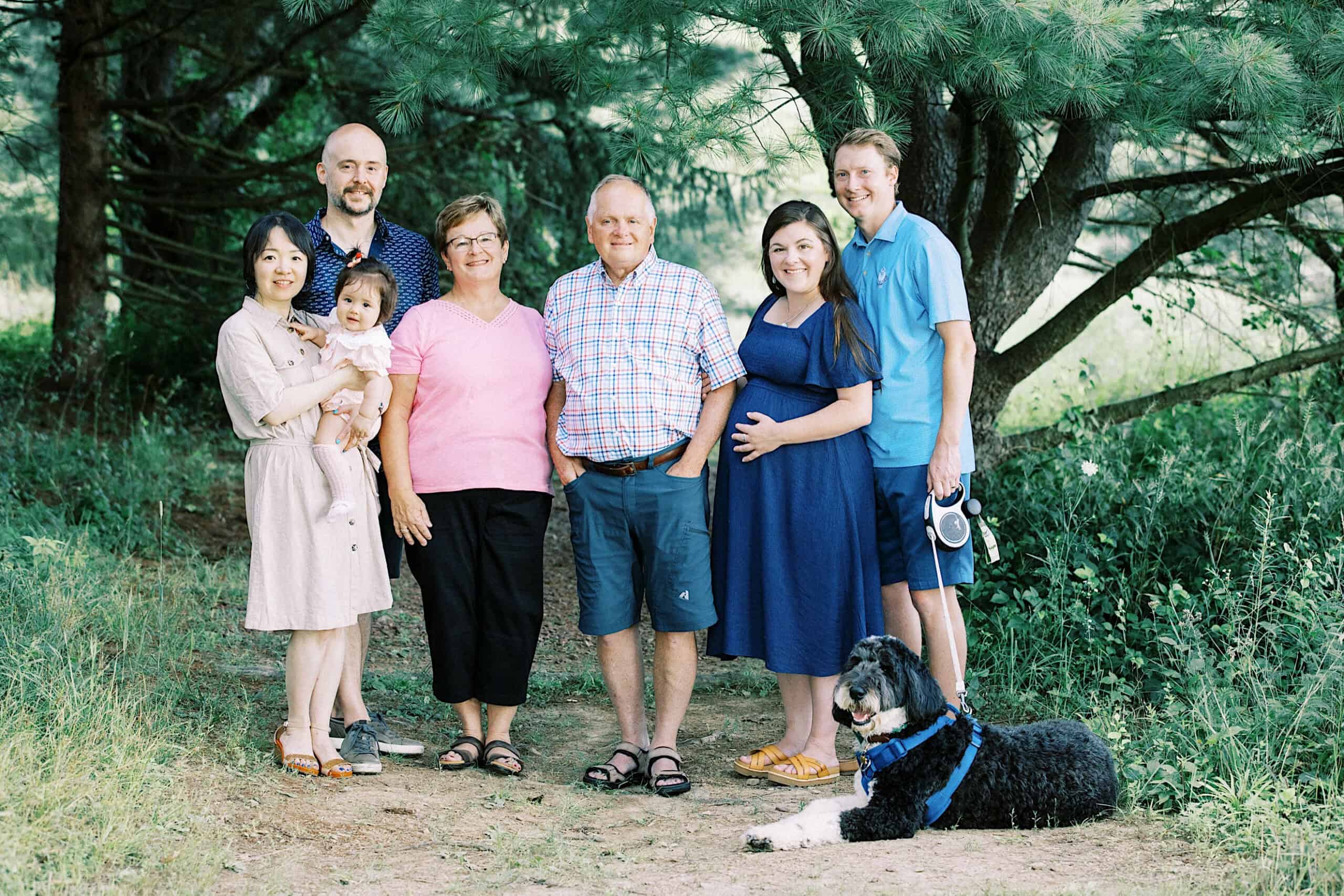 A group of seven adults and one child pose outdoors in a wooded area for extended family photos, with a black and white dog lying on the ground in front of them.
