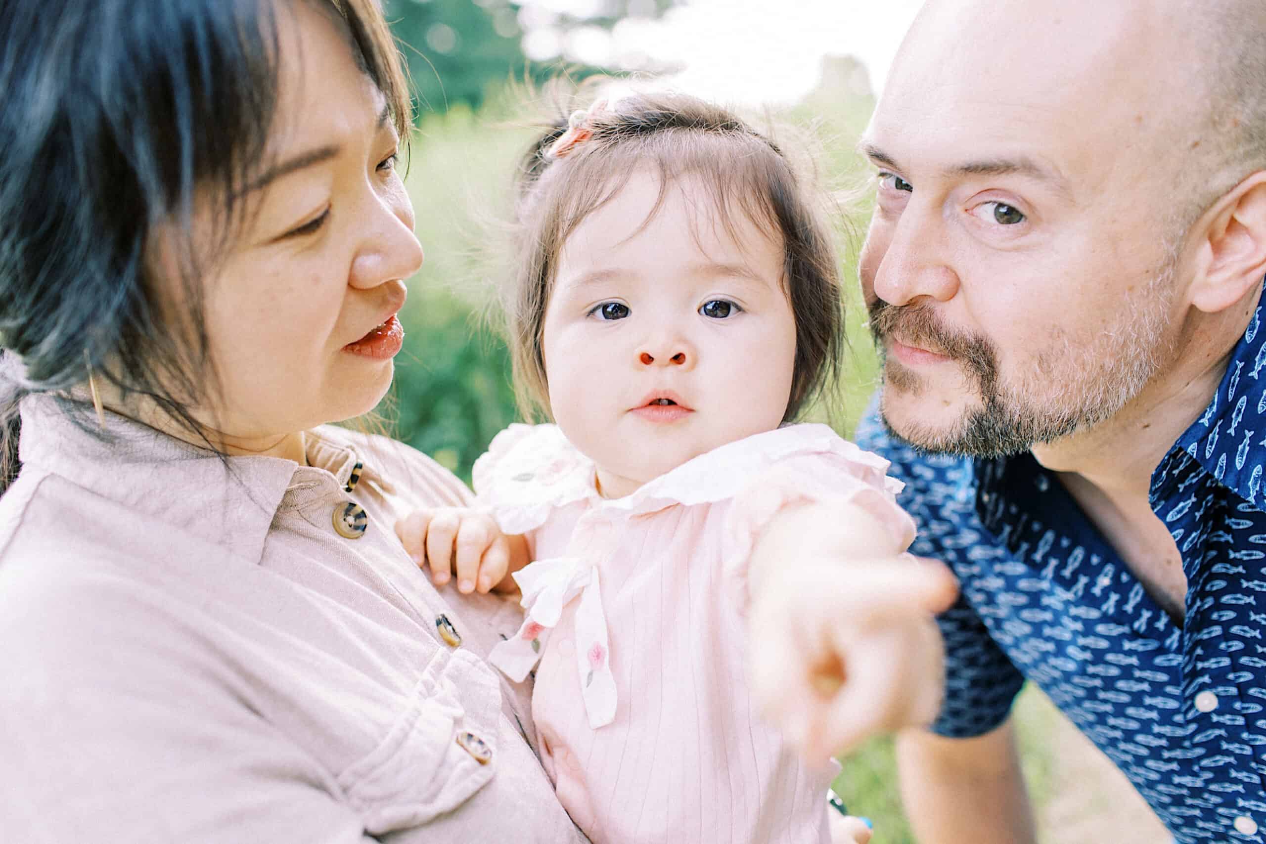 A woman holds a young child in a pink outfit while a man leans in close; the child points forward with a neutral expression, capturing the warmth often seen in maternity and extended family photos.