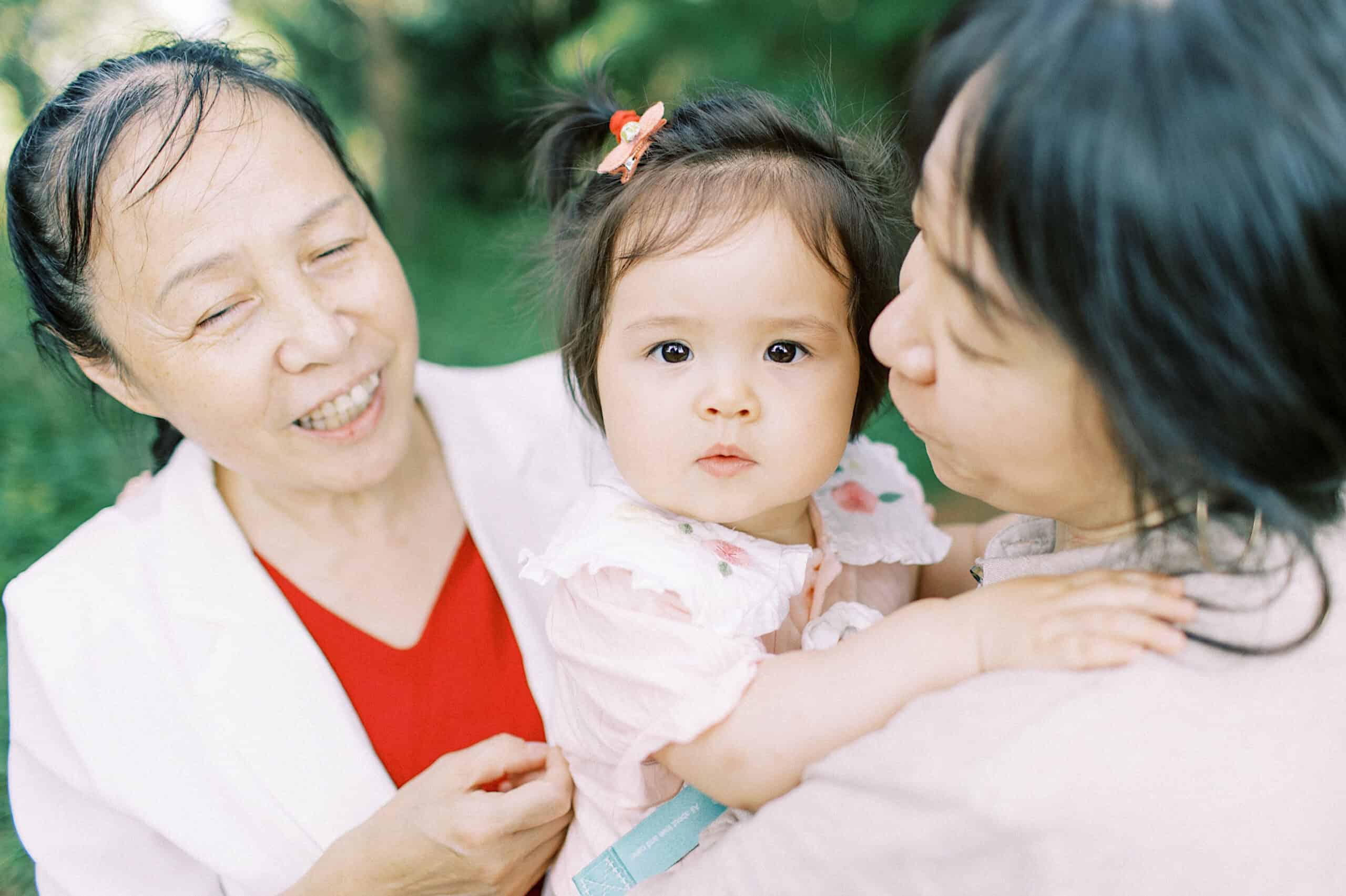 An elderly woman, a baby girl, and another woman are outdoors enjoying extended family photos. The baby is being held and looks at the camera as the women smile warmly, with green foliage in the background.