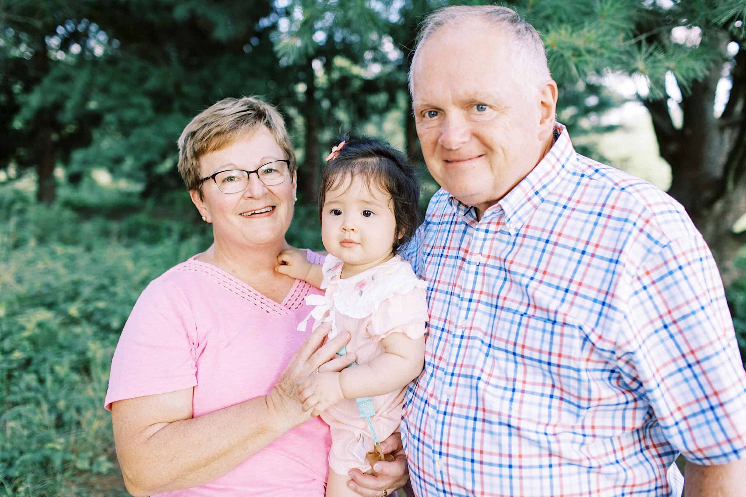 An older woman and man stand outdoors, smiling and holding a young child between them, capturing a heartwarming moment perfect for extended family photos. Trees and greenery are visible in the background.