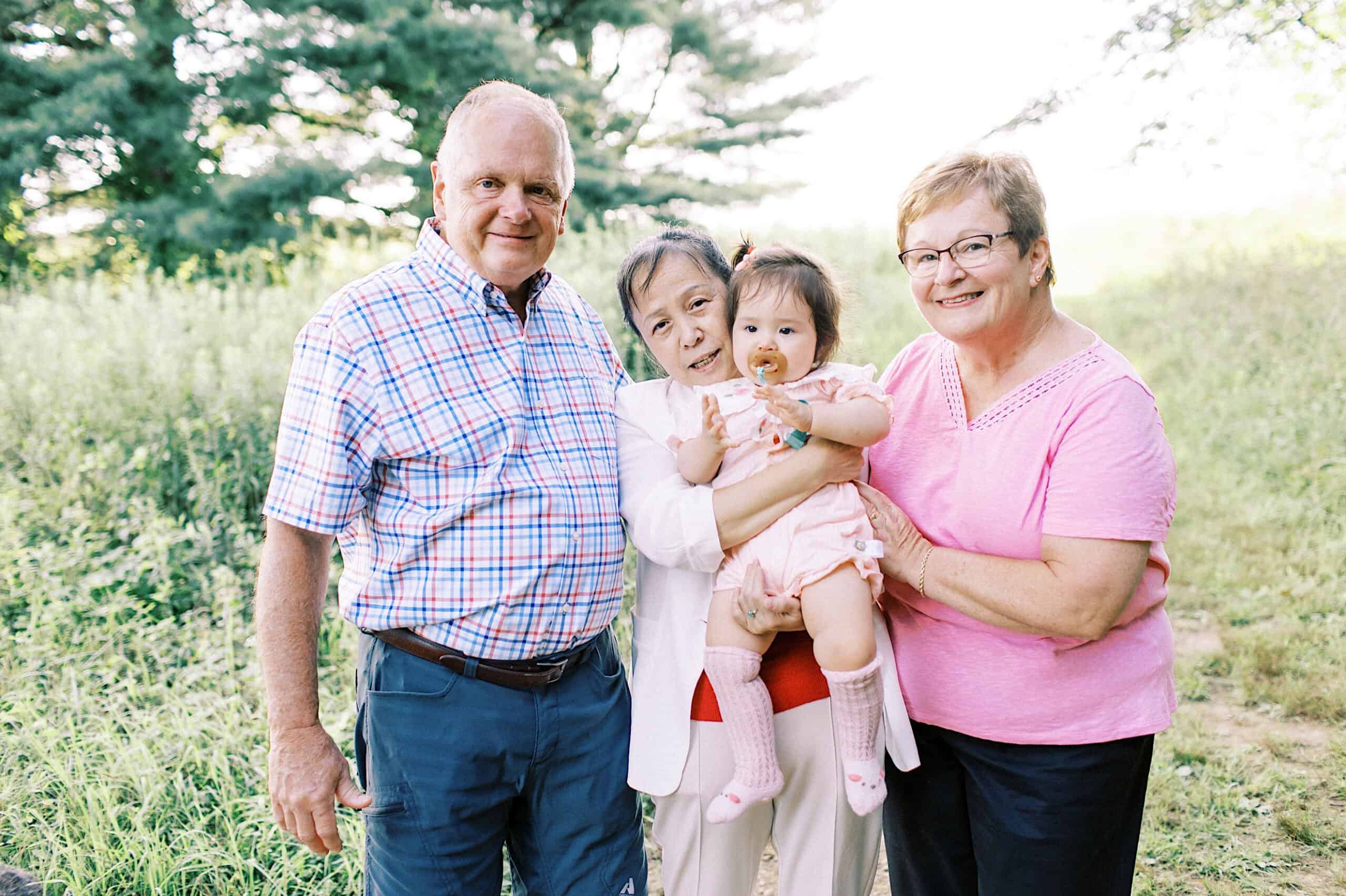 Three adults stand outdoors with a baby in their arms on a sunny day, surrounded by greenery—capturing the joy of maternity and extended family photos.