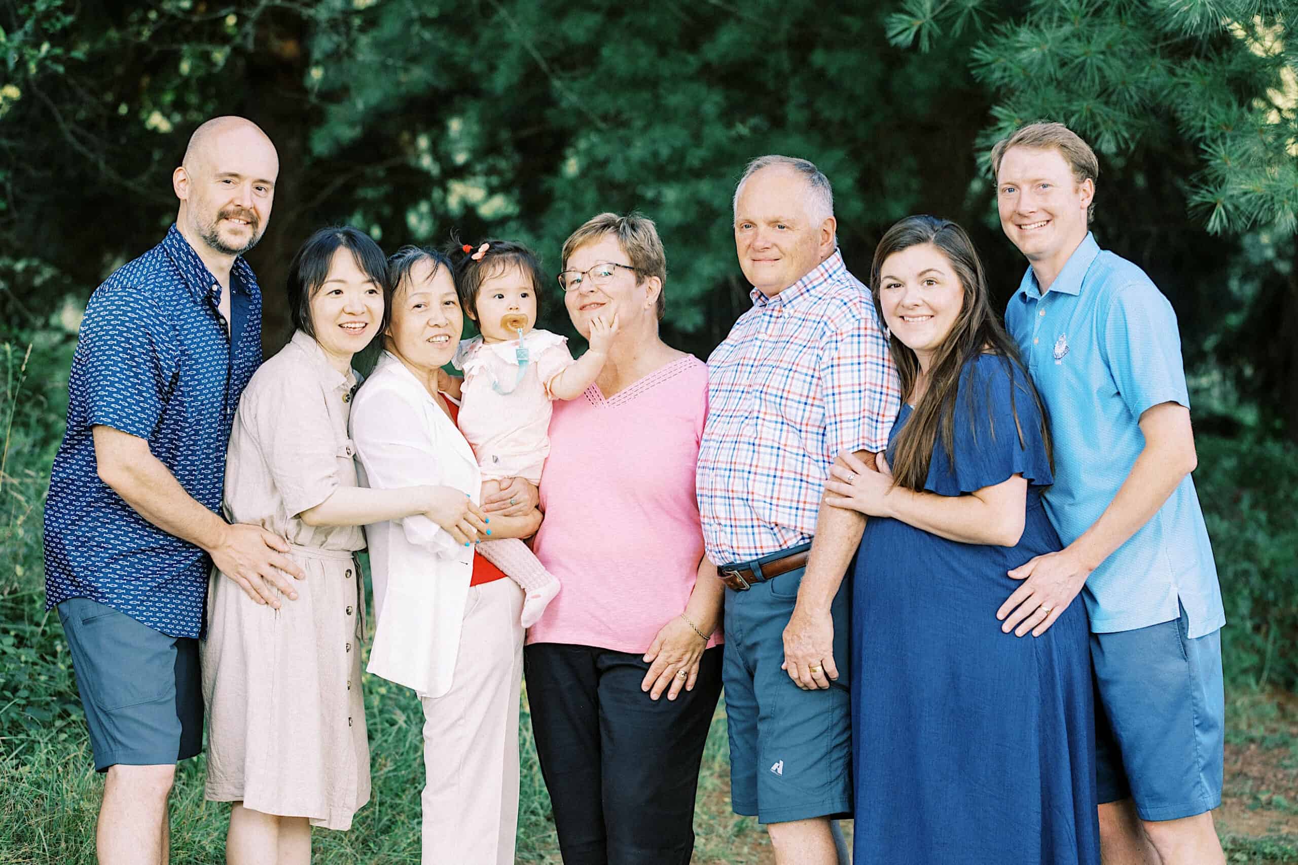 Eight adults and one young child stand outdoors in front of green trees, posing closely together and smiling at the camera—an ideal moment for maternity and extended family photos.