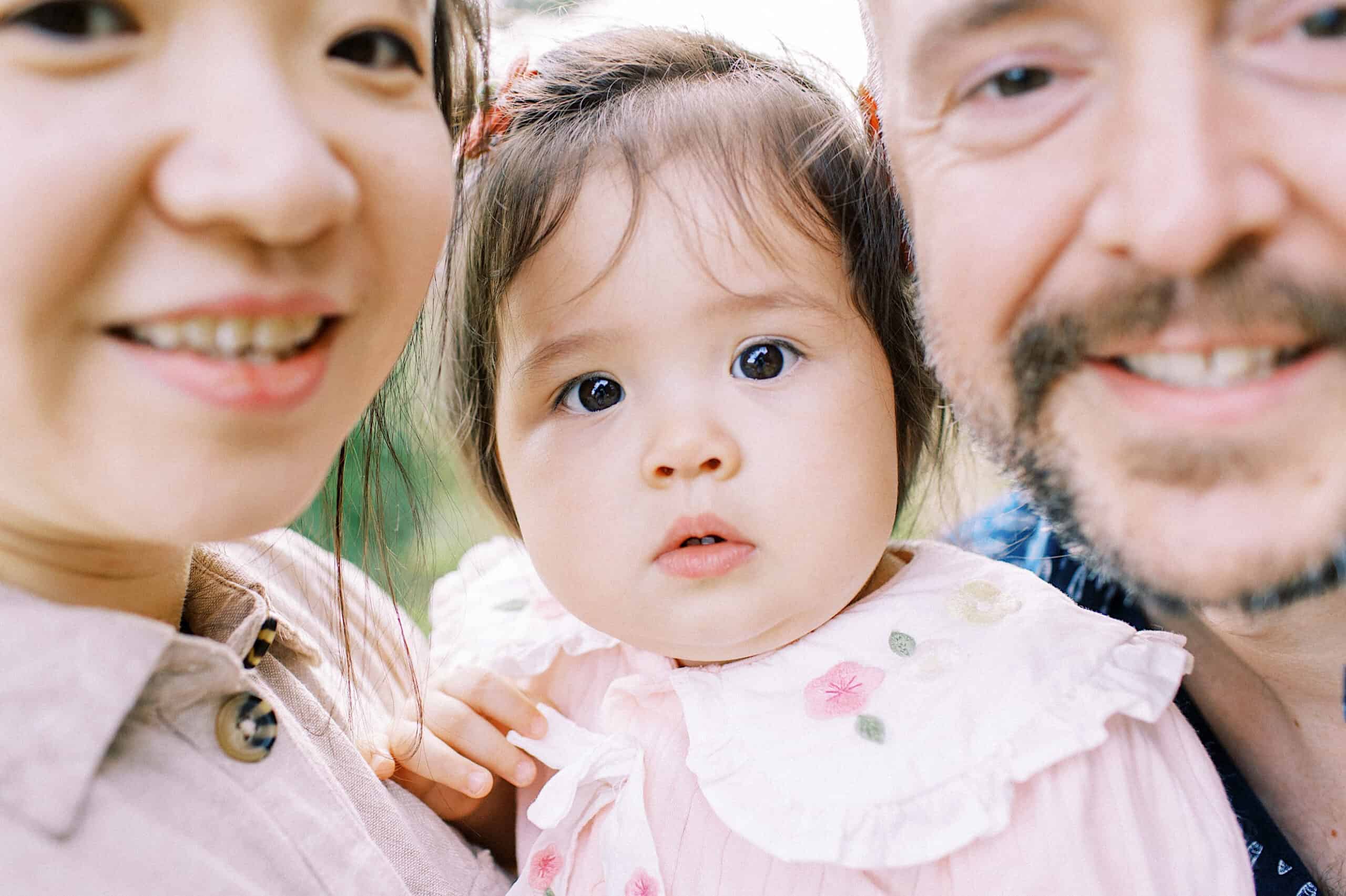 Close-up of two adults and a baby looking at the camera outdoors, perfect for maternity and extended family photos. The baby is in the center, wearing a light pink outfit with embroidery.