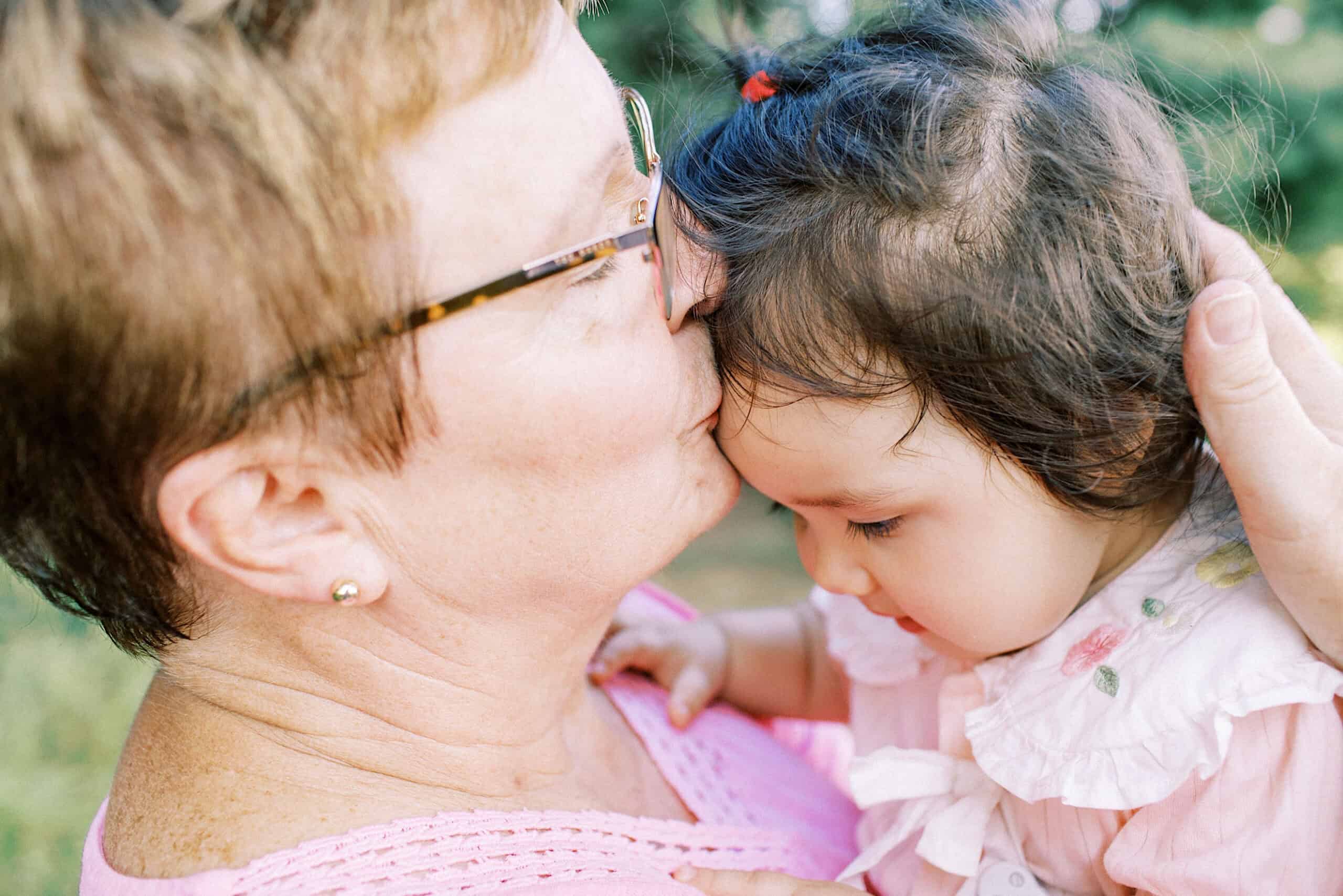 An older woman wearing glasses kisses a young child on the forehead while holding her, both dressed in light pink clothing outdoors—capturing a tender moment perfect for maternity and extended family photos.