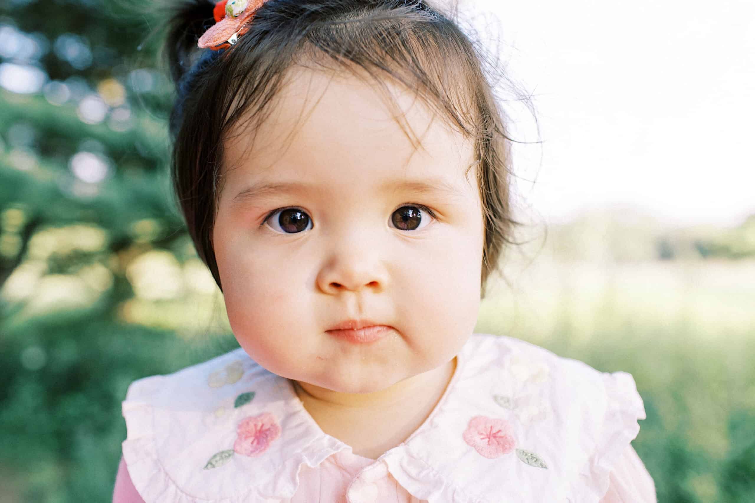 A young child with dark hair and a floral embroidered shirt looks directly at the camera outdoors, surrounded by greenery—perfect for capturing candid moments during maternity and extended family photos.
