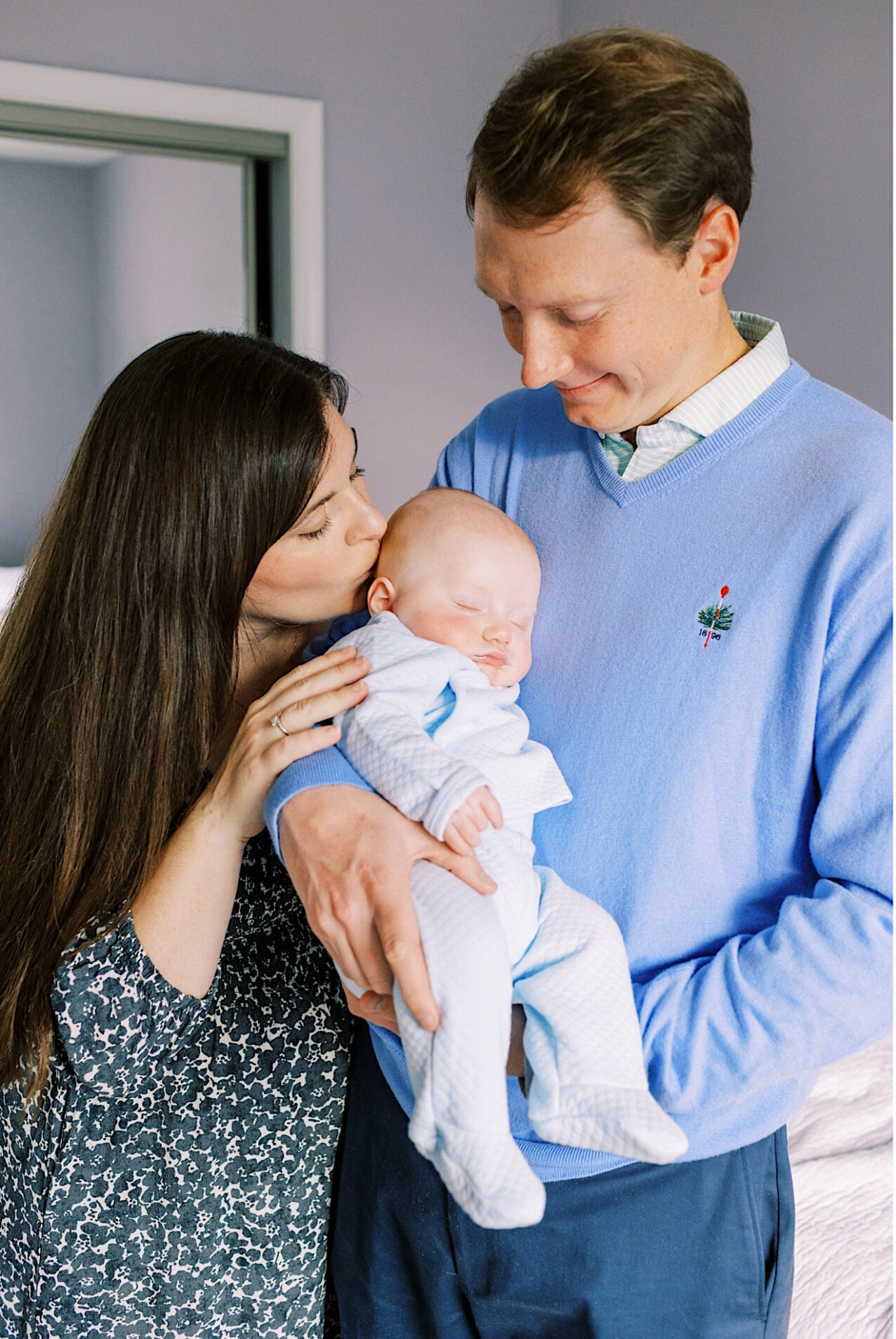 A man holds a sleeping baby dressed in light blue, while a woman kisses the baby's head. Capturing tender lifestyle at home newborn photos with soft indoor lighting sets a warm, intimate scene.