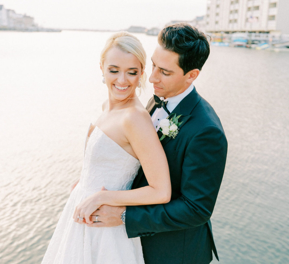 A bride and groom stand close together by the water at Stone Harbor, with the groom looking at the bride and the bride smiling.