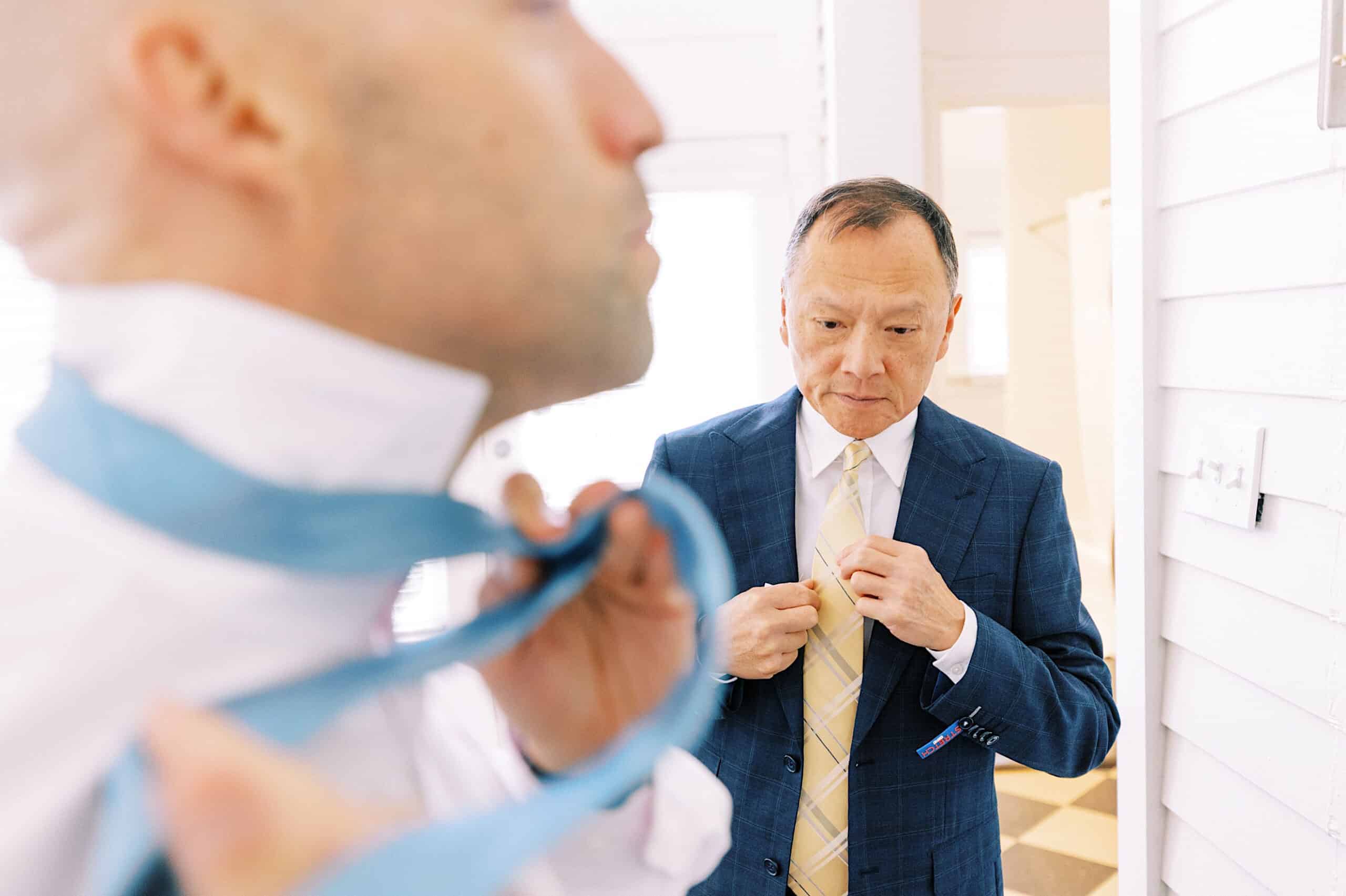 Two men in suits stand indoors at a Cape May wedding at the Virginia Hotel; one in the foreground adjusts a blue tie, while the other in the background fastens a yellow tie and looks down.