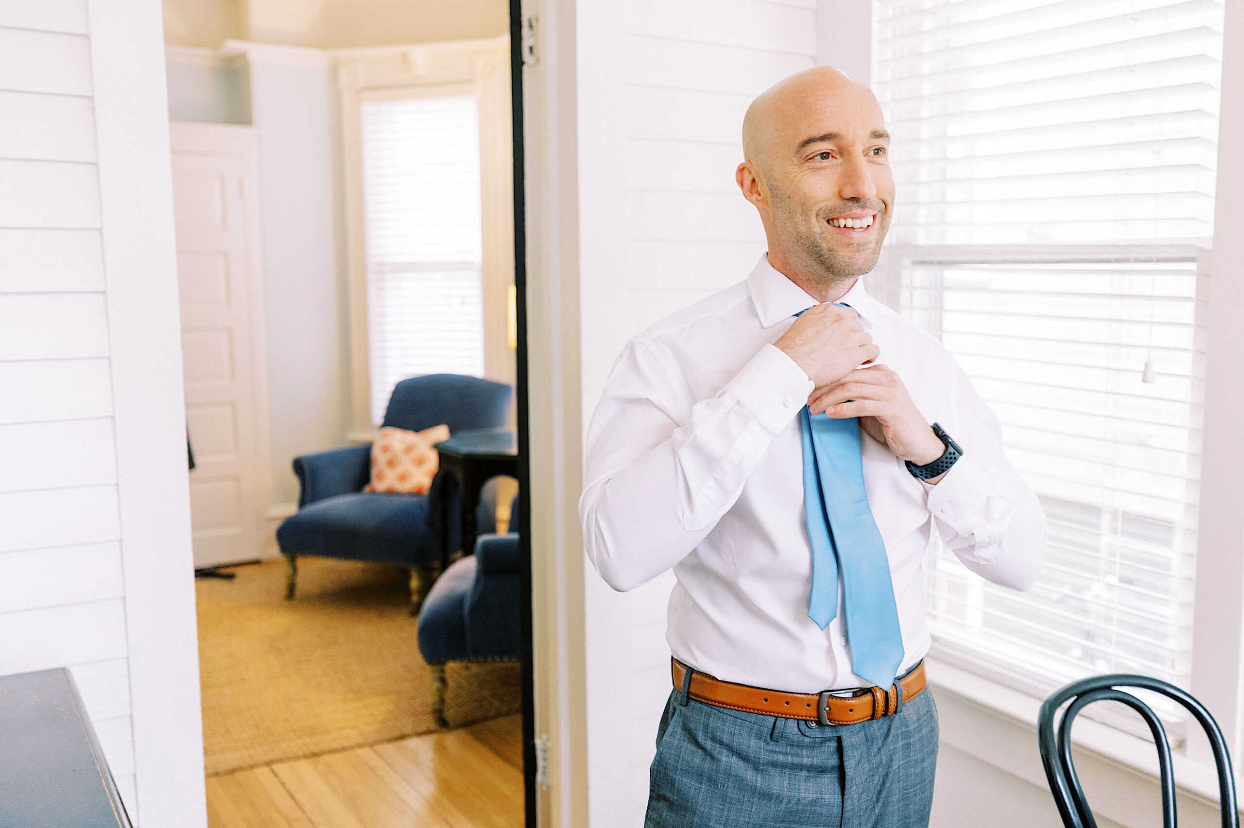 A man in a white shirt and blue tie stands in a bright room, adjusting his tie and smiling, ready for a Cape May wedding at the Virginia Hotel. A blue chair and window blinds are visible in the background.
