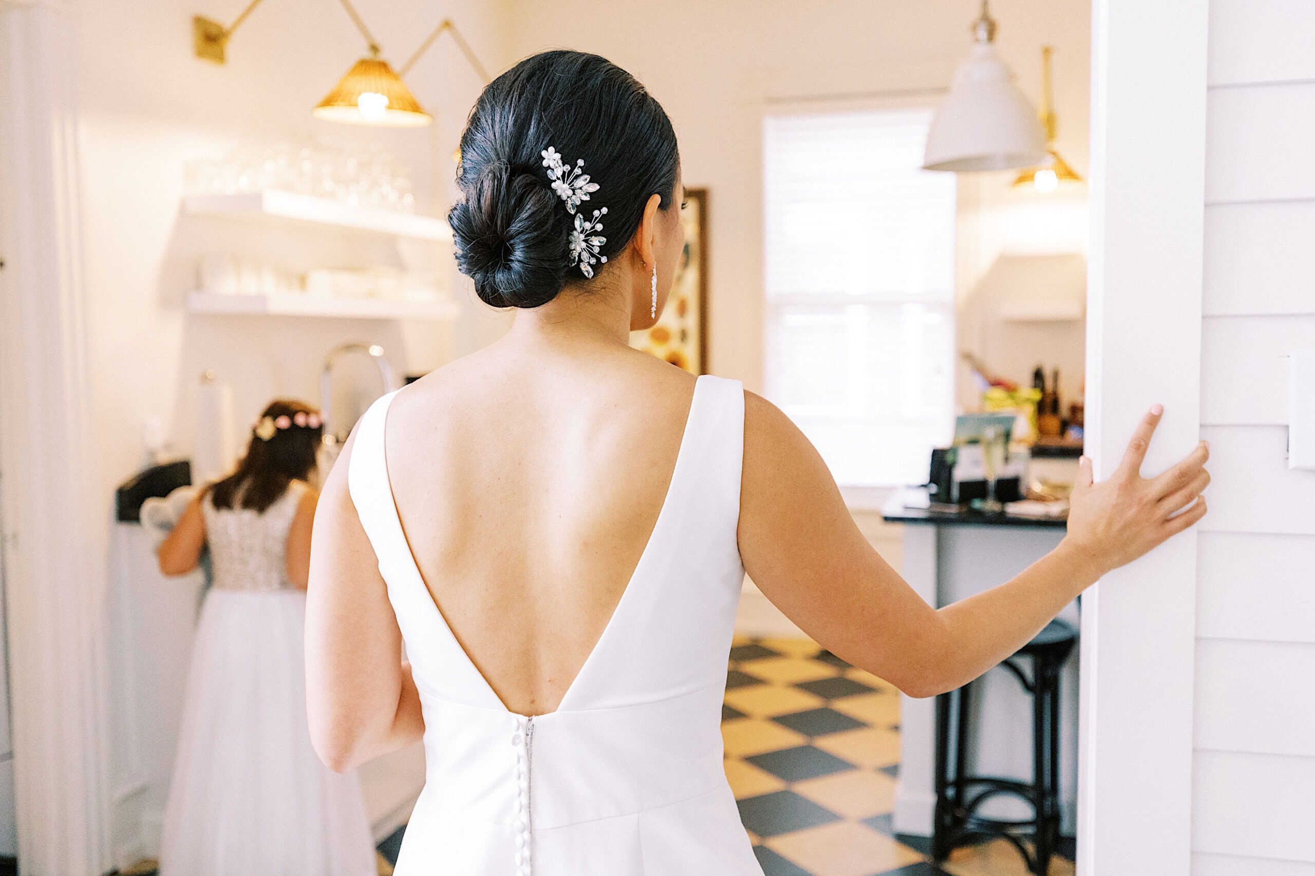 A woman in a white dress with a low back and decorative hairpin stands in a bright room at the Virginia Hotel, looking toward a girl in a white dress—capturing the timeless elegance of a Cape May wedding.