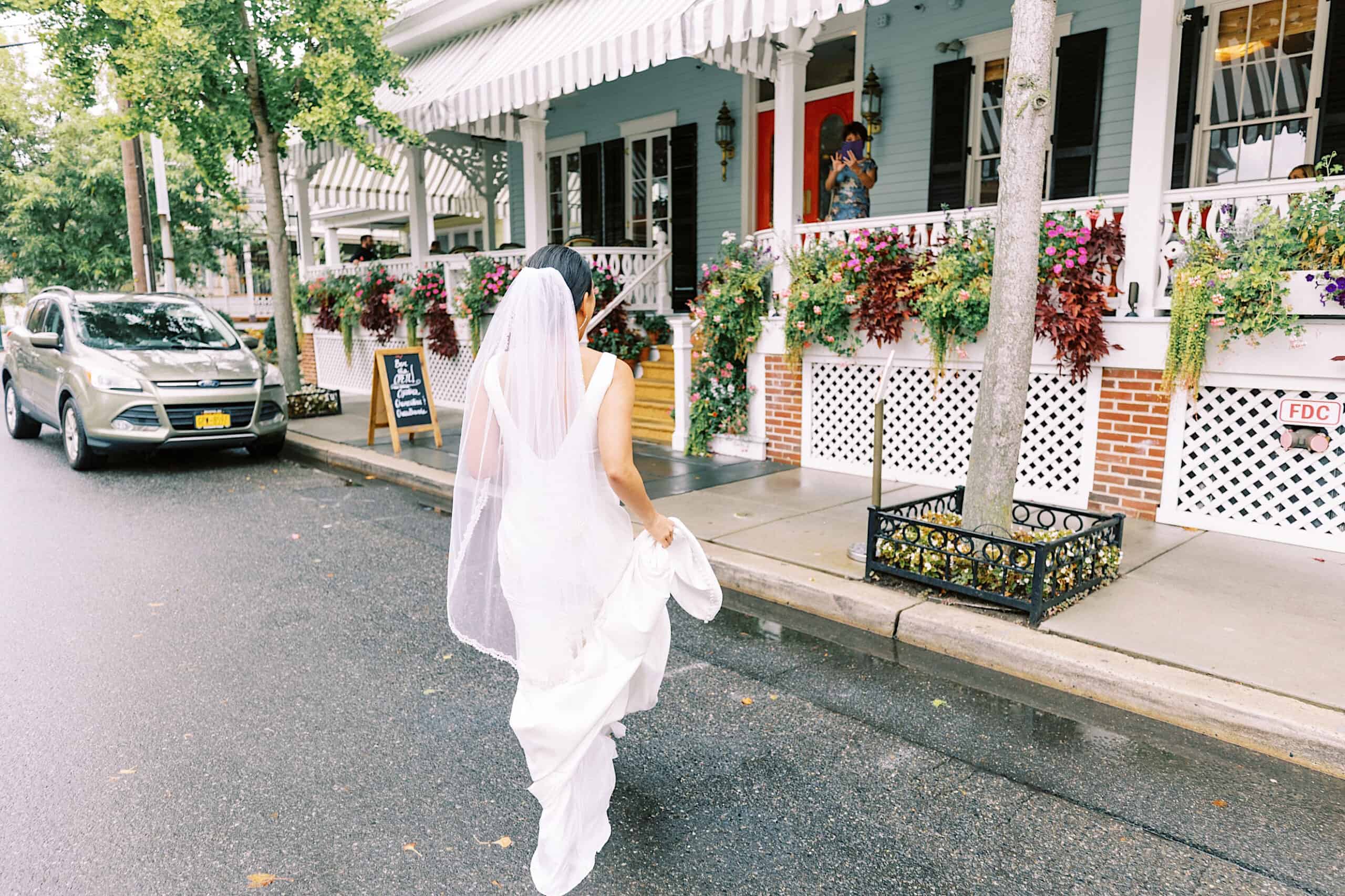 A bride in a white dress and veil walks along a wet street toward the flower-adorned porch, capturing the romance of a Cape May wedding at the Virginia Hotel.