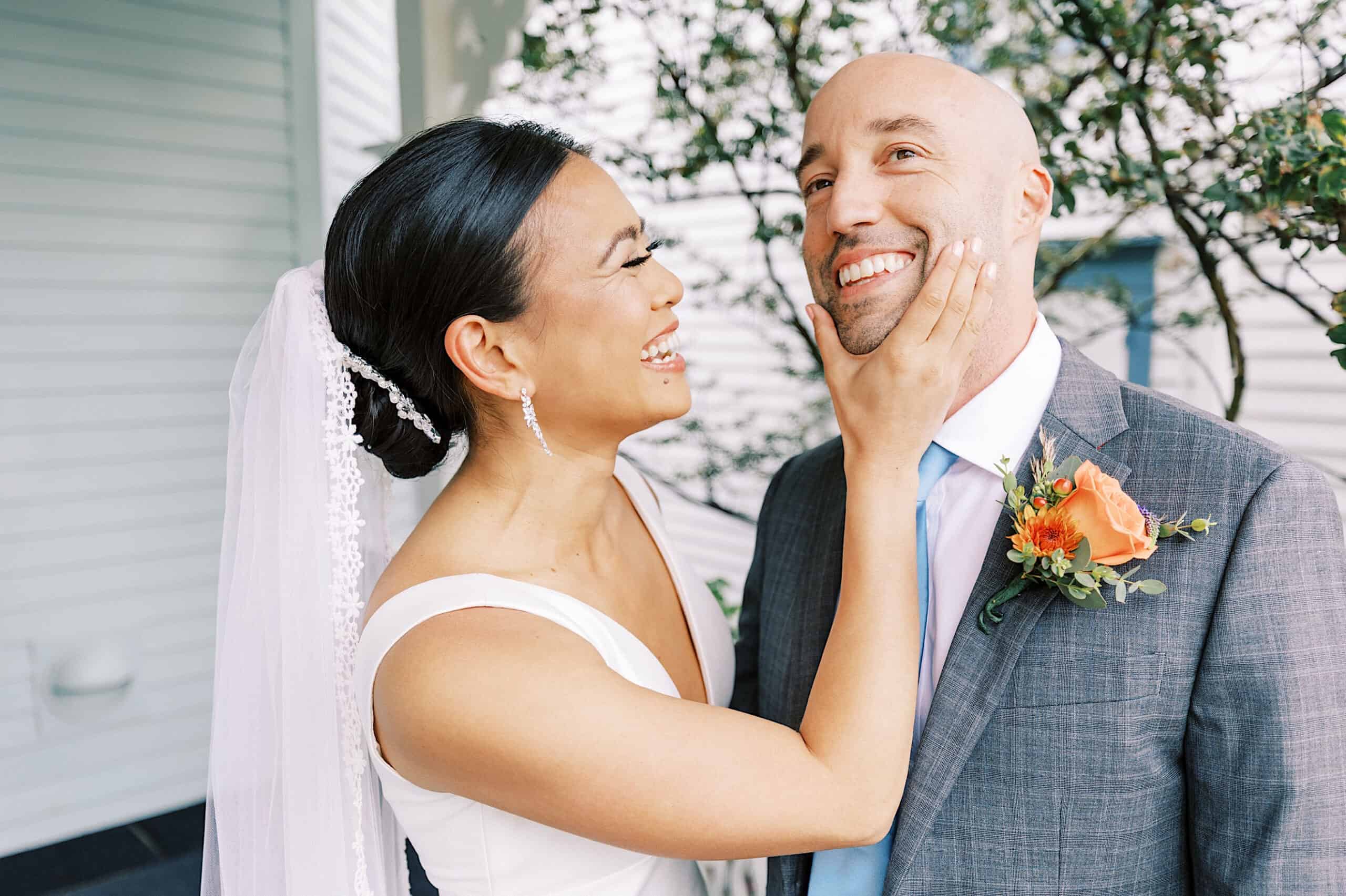 A bride in a white dress and veil smiles while holding the groom's face during their Cape May wedding at the Virginia Hotel; the groom, in a gray suit with an orange boutonniere, smiles back.