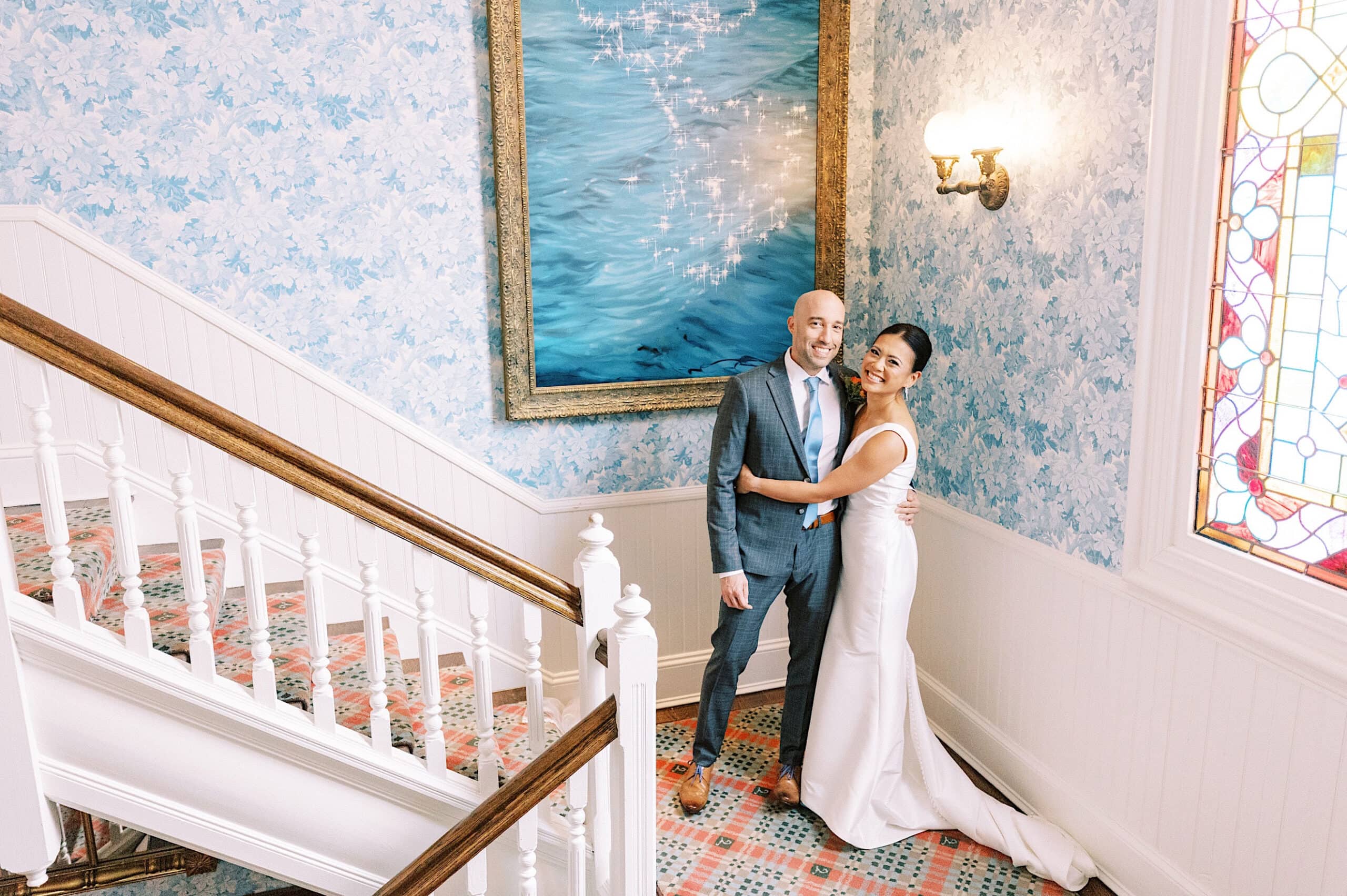 A couple poses together on a staircase in formal wedding attire at their Cape May wedding at the Virginia Hotel, with light blue patterned walls and a large painting behind them.