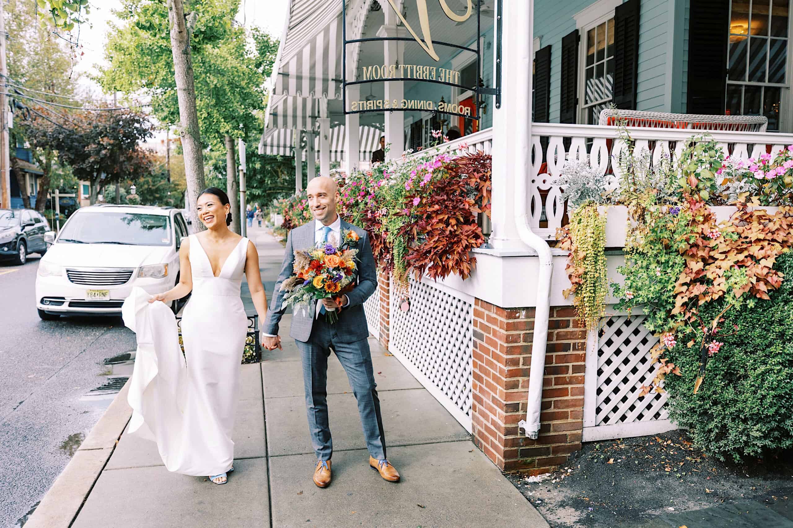A bride and groom walk hand in hand on a sidewalk outside the flower-adorned Virginia Hotel, both smiling during their beautiful Cape May wedding.