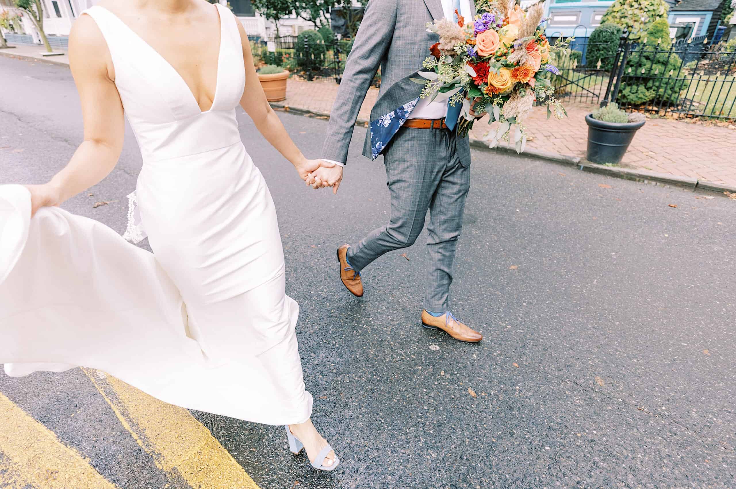 A bride in a white dress and a groom in a gray suit holding a large bouquet walk hand-in-hand down a wet street, capturing the romantic charm of a Cape May wedding at the Virginia Hotel.
