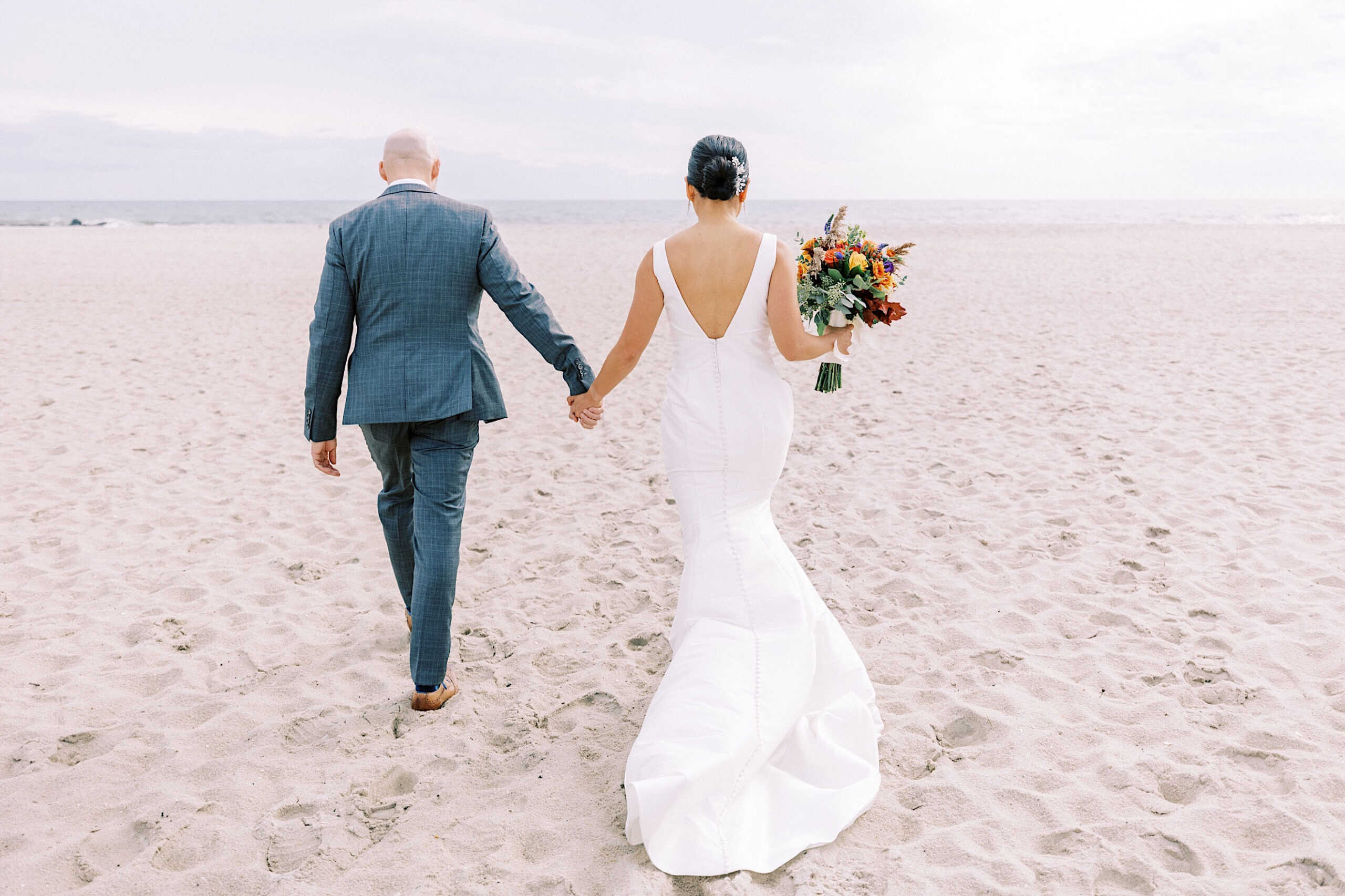A bride in a white dress and a groom in a blue suit walk hand in hand on a sandy beach toward the ocean, capturing the romance of a Cape May wedding at the Virginia Hotel.