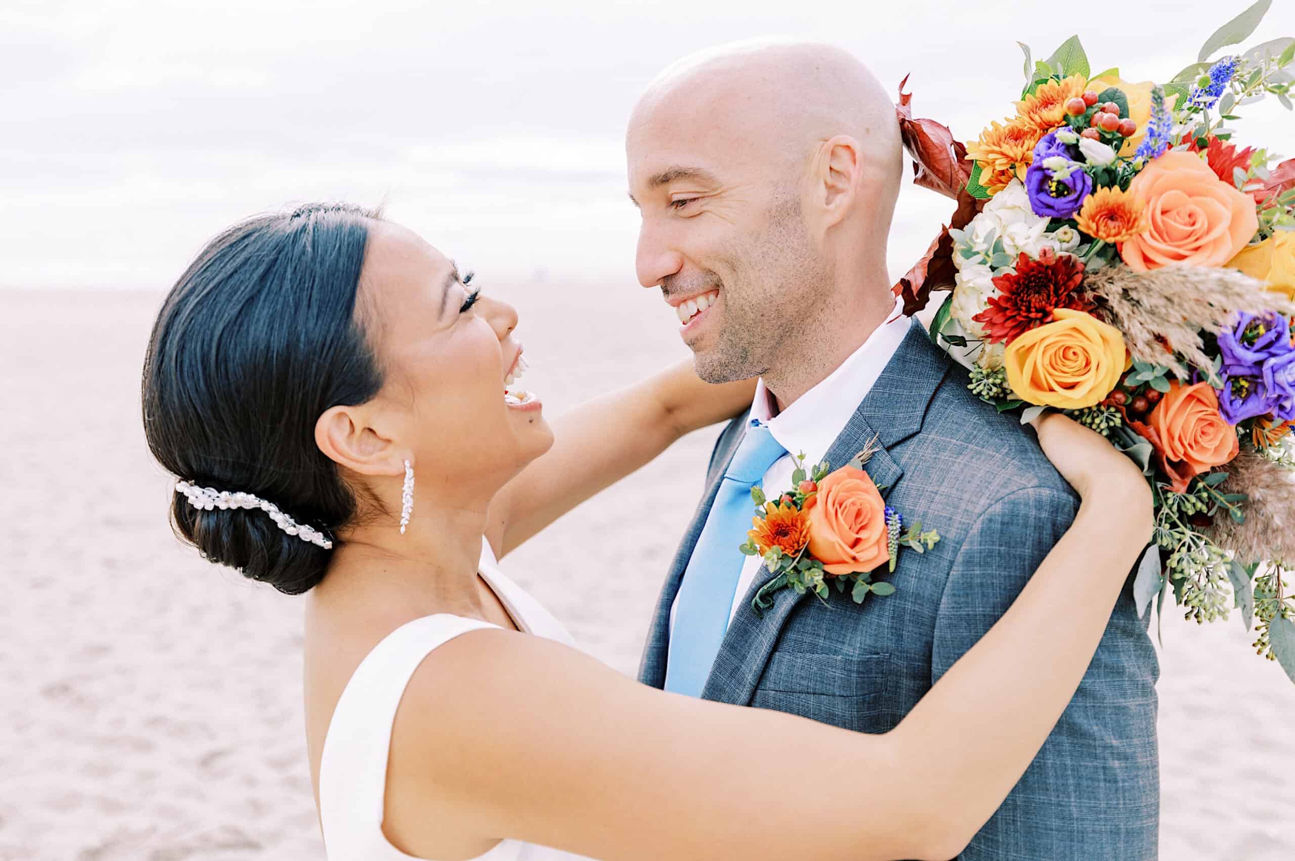 A bride and groom stand on a beach, smiling at each other during their Cape May wedding at the Virginia Hotel. The bride holds a colorful bouquet, her arms wrapped lovingly around the groom's shoulders.