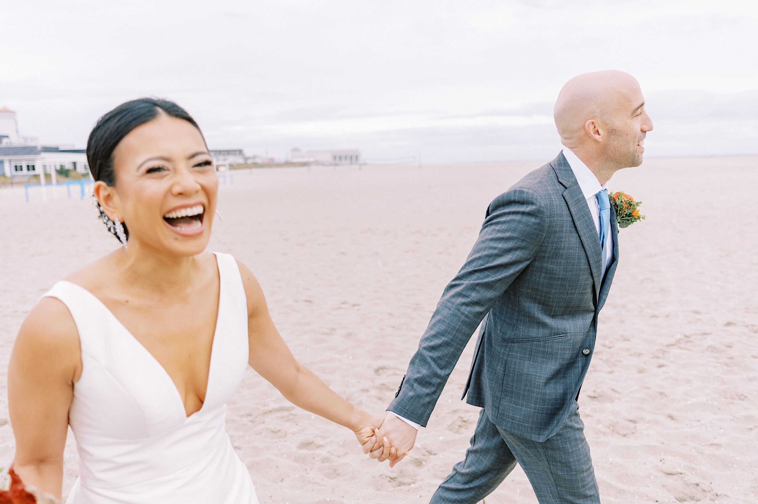 A smiling bride in a white dress and a groom in a gray suit walk hand-in-hand on a sandy beach during their Cape May wedding at the Virginia Hotel.