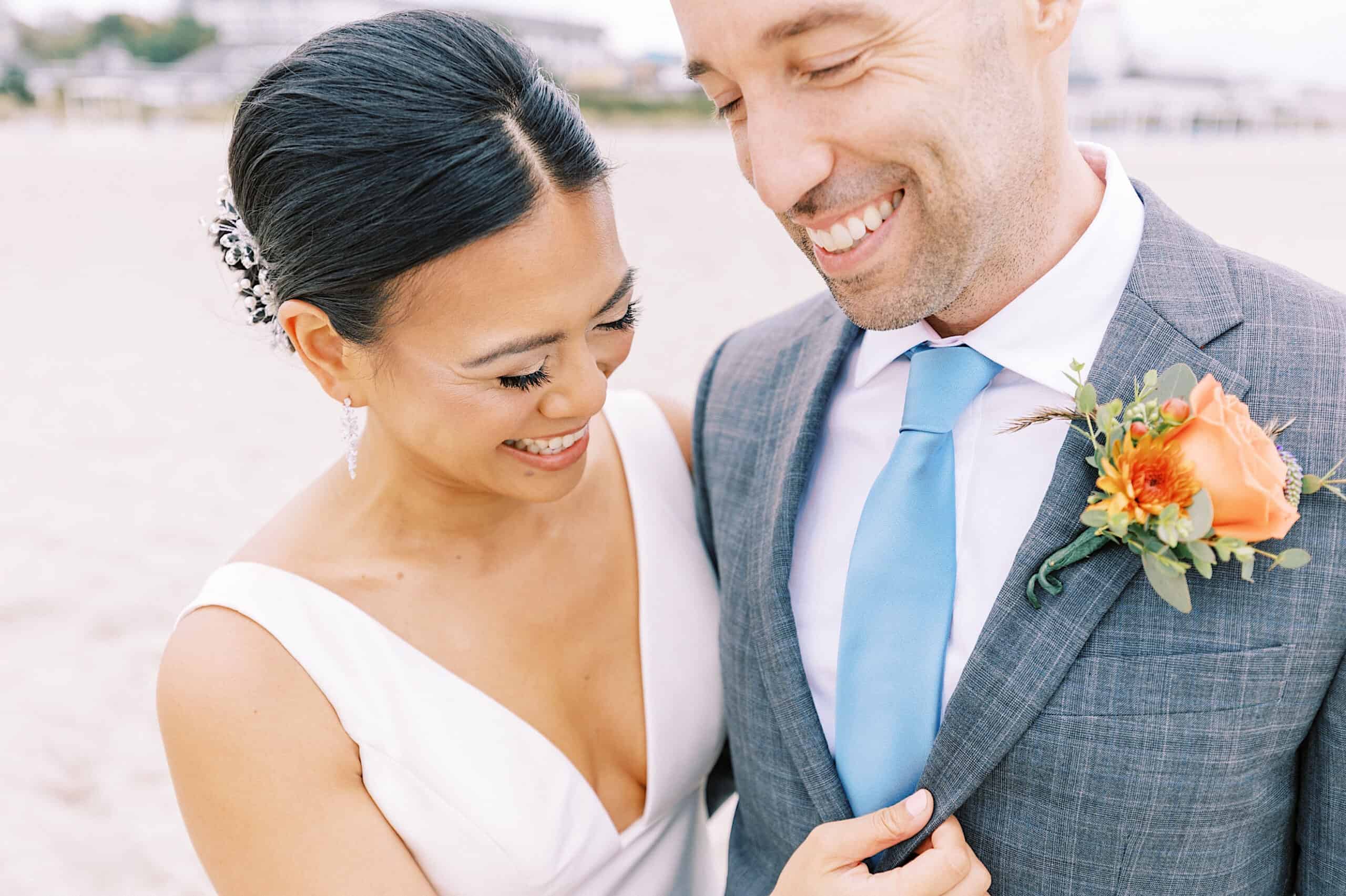 A bride and groom stand together outdoors, smiling, at their Cape May wedding at the Virginia Hotel; the groom wears a gray suit with a blue tie and orange boutonniere, and the bride wears a white dress.