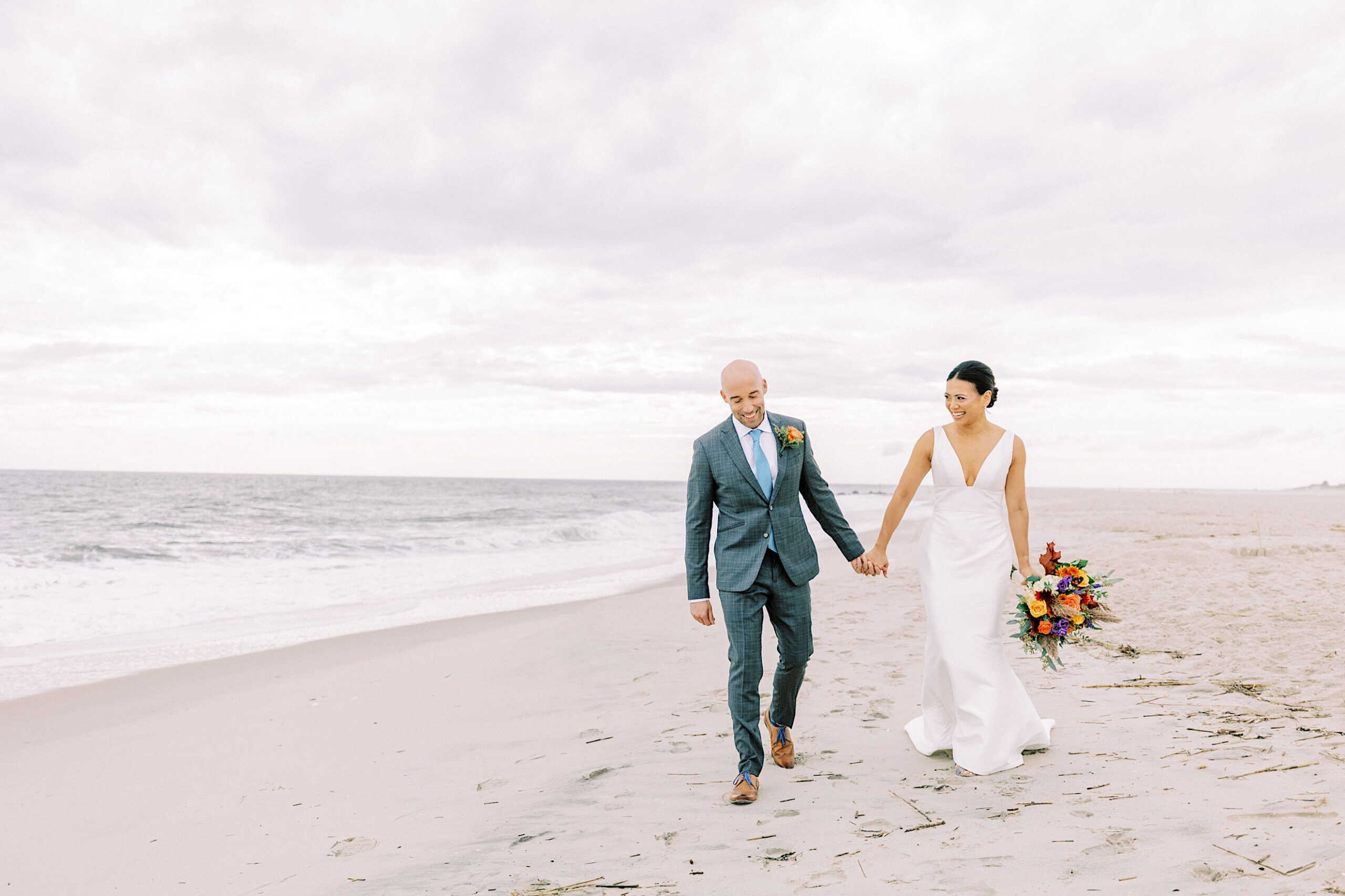A couple dressed in wedding attire walks hand in hand along a sandy beach at their elegant Cape May wedding at the Virginia Hotel, with the ocean and cloudy sky as a backdrop. The bride holds a colorful bouquet.