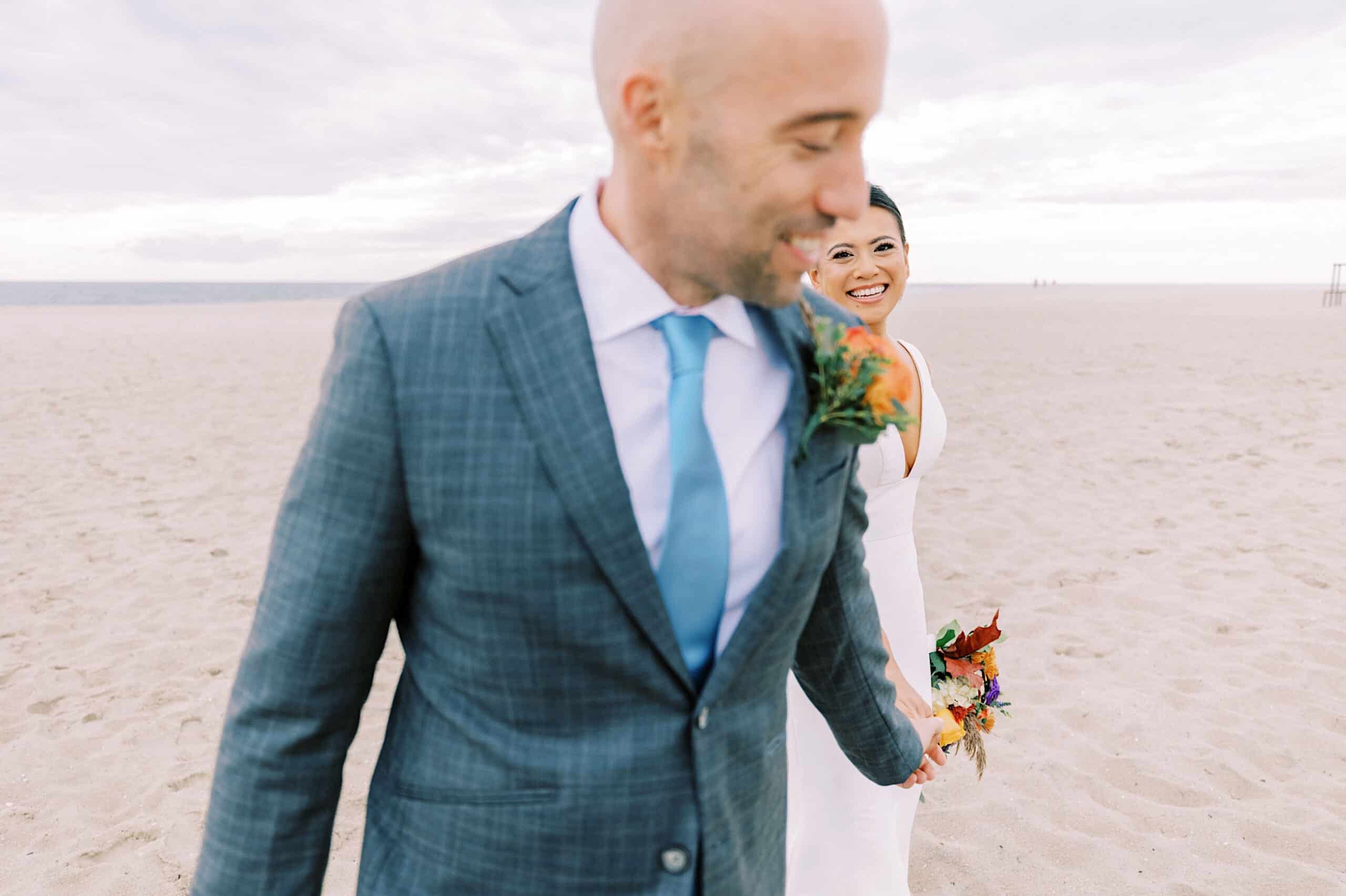 A man in a suit leads a woman in a white dress holding a bouquet across a sandy beach, capturing the romance of a Cape May wedding at the Virginia Hotel.