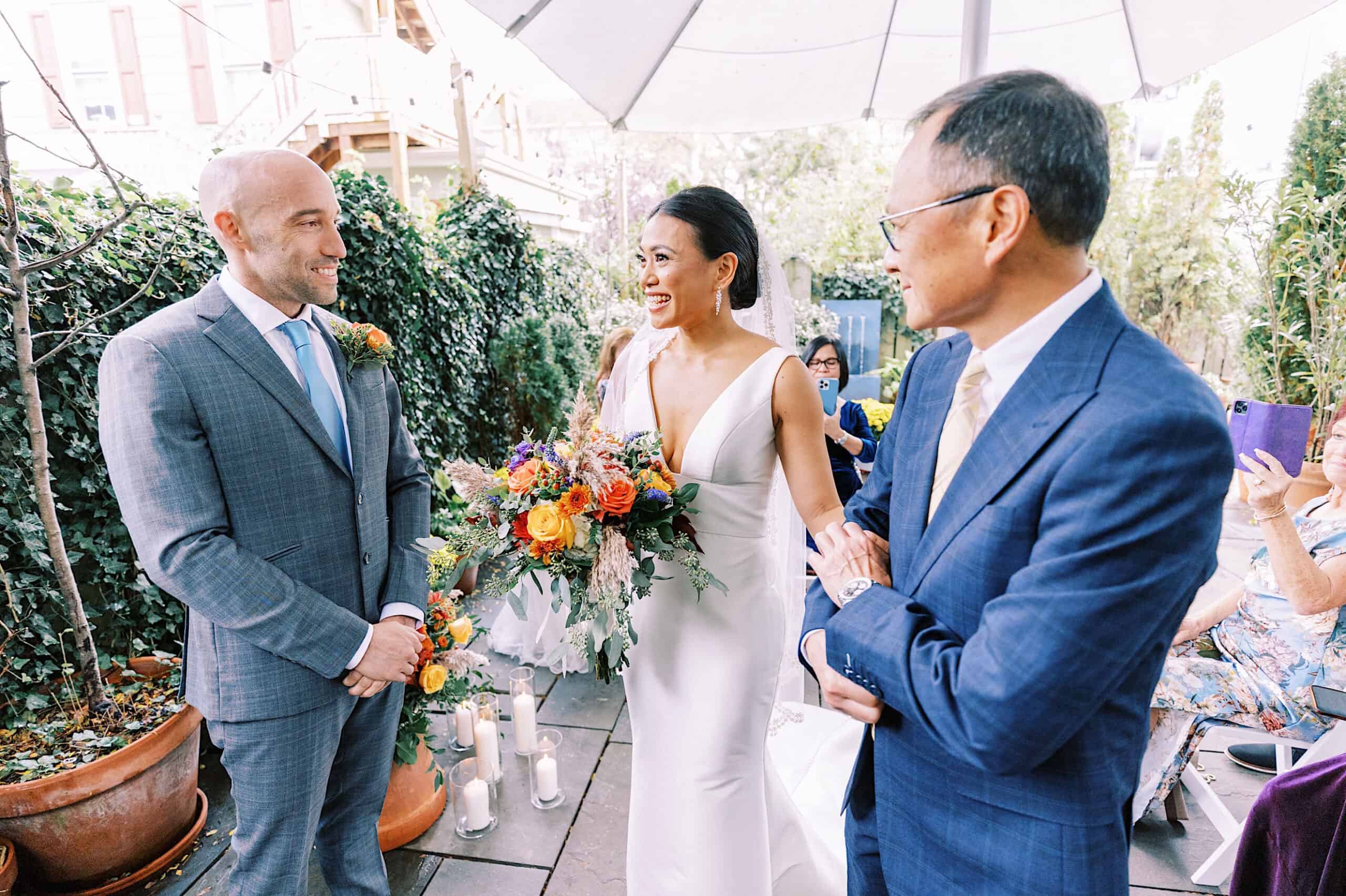 A bride holding a colorful bouquet stands between two men in suits at an outdoor Cape May wedding at the Virginia Hotel, with guests seated and lush greenery in the background.