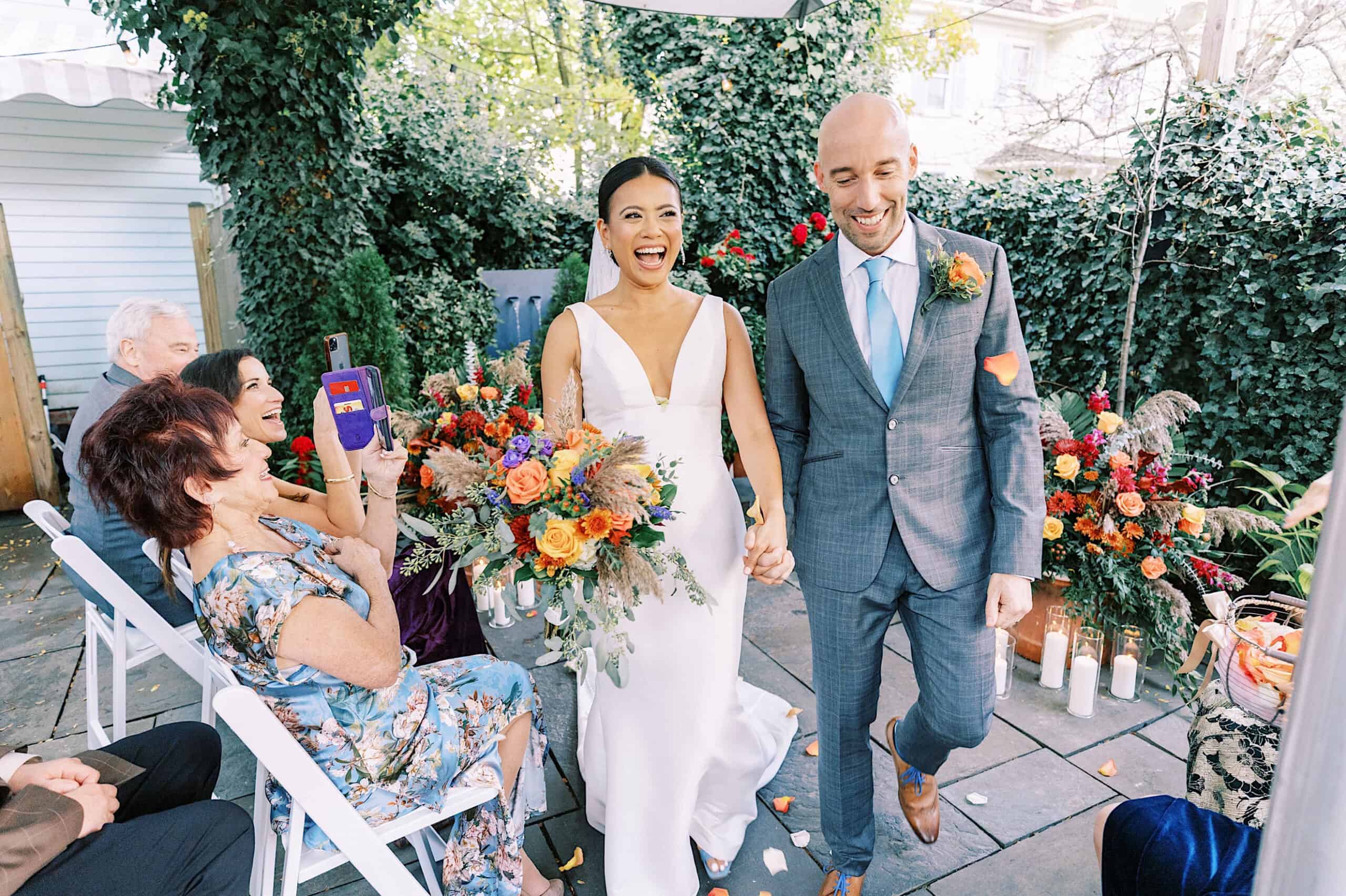 A bride and groom walk down the aisle holding hands, smiling, surrounded by seated guests and colorful floral arrangements at their Cape May wedding at the Virginia Hotel.