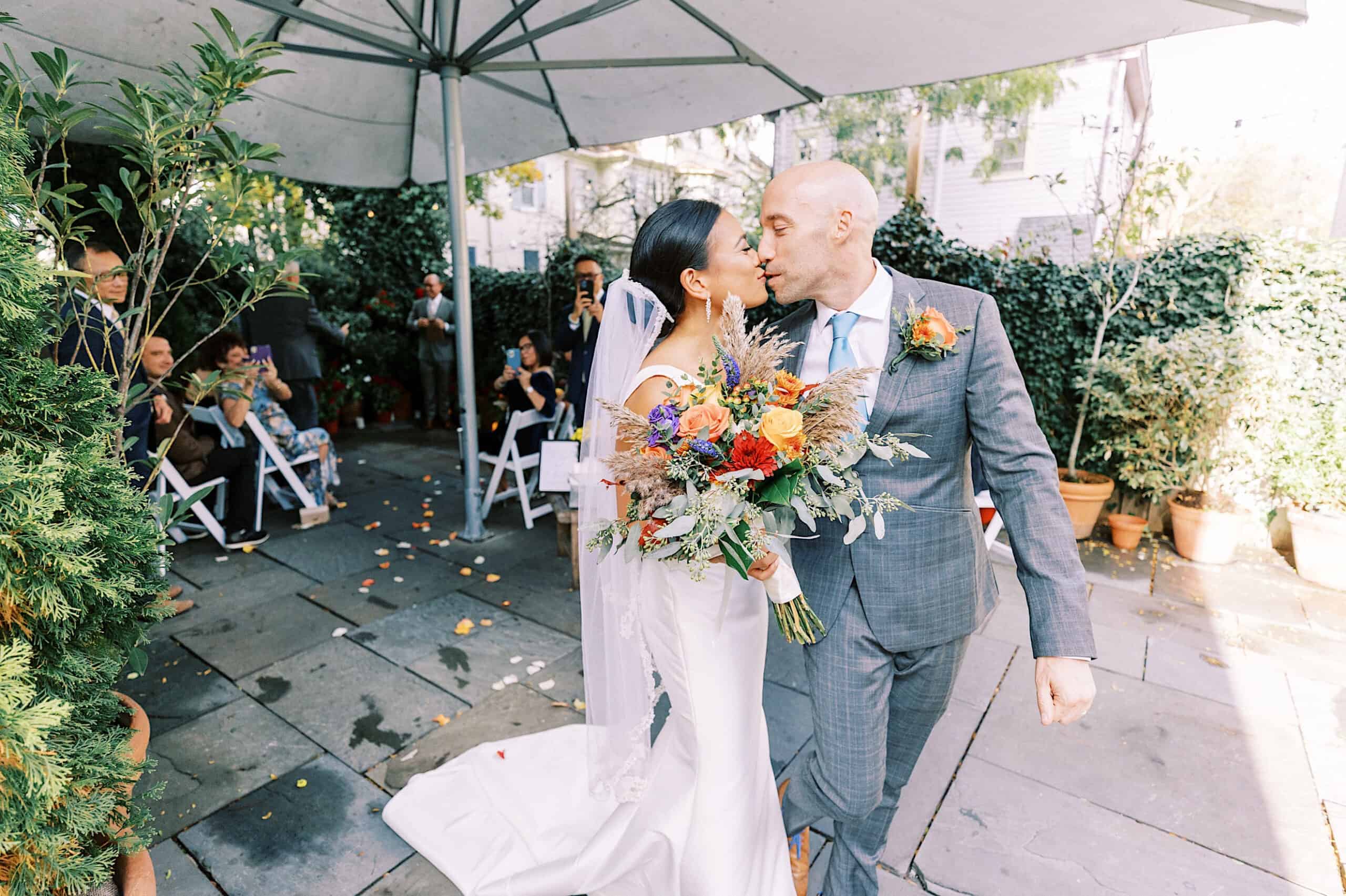 A bride and groom kiss while walking outdoors at their Cape May wedding at the Virginia Hotel, holding a bouquet, with guests seated and lush greenery in the background.