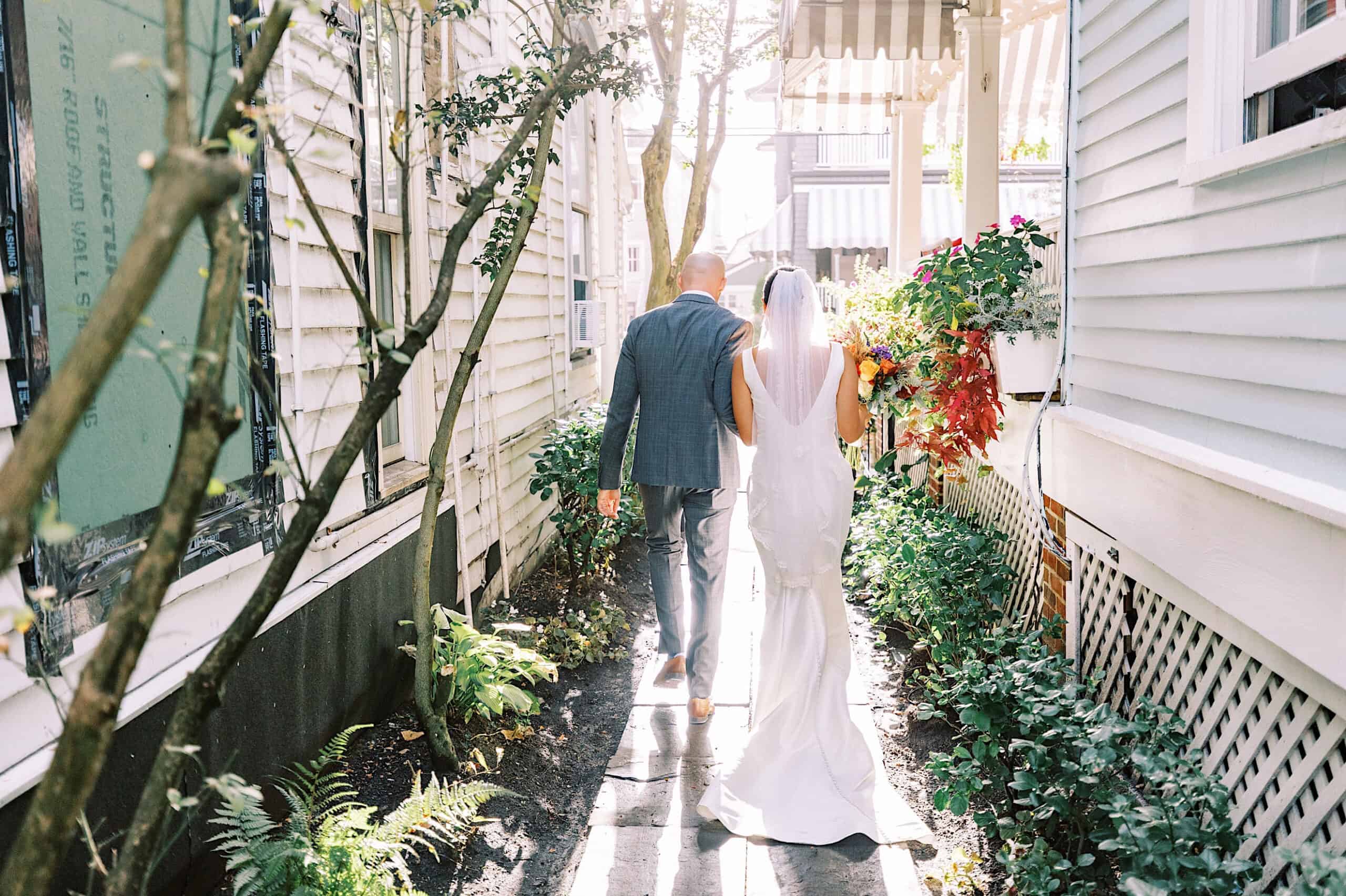 A bride in a white gown and veil walks beside a man in a suit down a sunlit, narrow outdoor pathway lined with greenery and white buildings during a Cape May wedding at the Virginia Hotel.