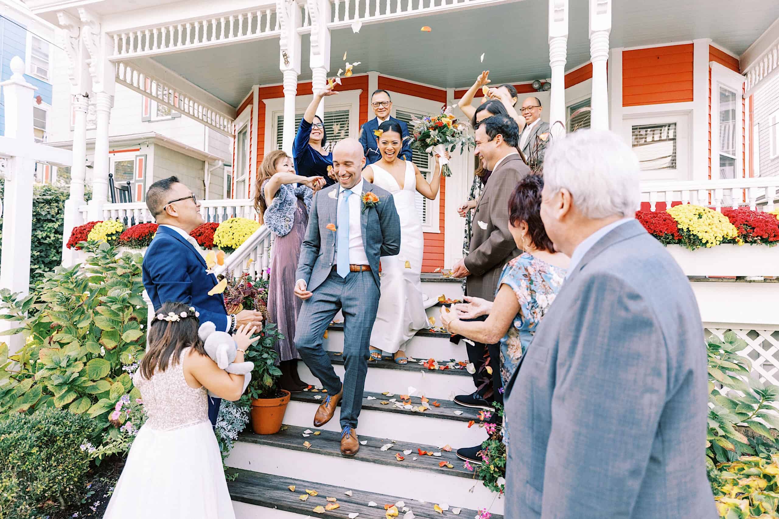 A newlywed couple walks down steps at their Cape May wedding at the Virginia Hotel, surrounded by smiling guests tossing flower petals in celebration.