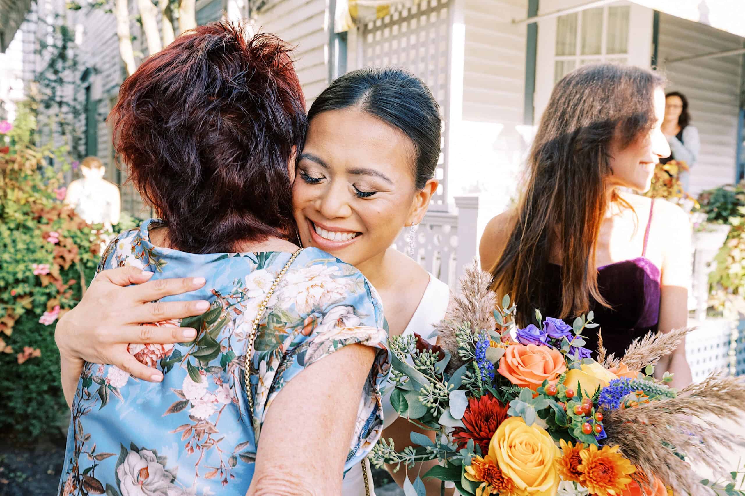 A woman in a white dress smiles while hugging another woman in a floral shirt, holding a colorful bouquet of flowers during a Cape May wedding at the Virginia Hotel. Another woman stands nearby.