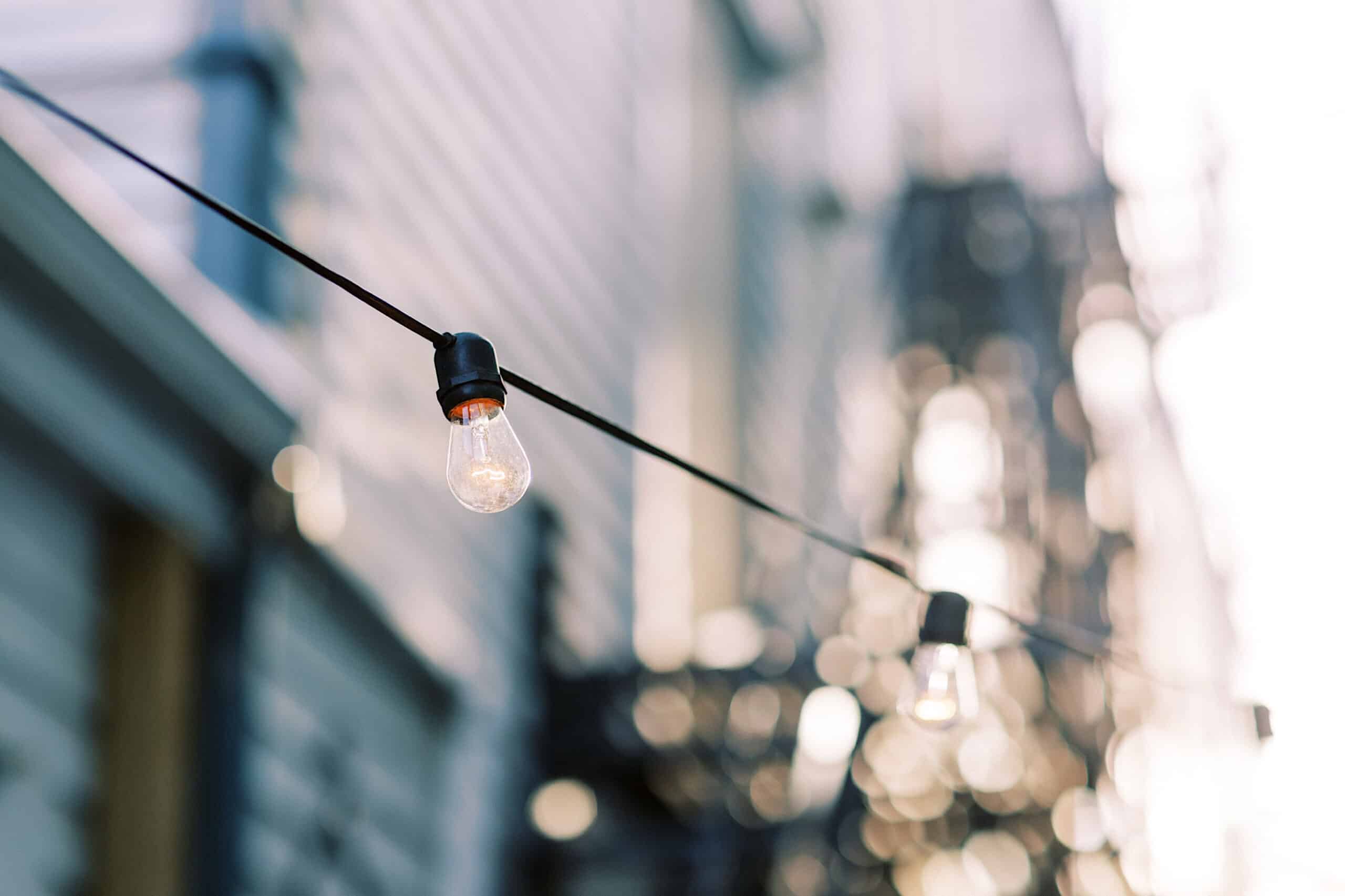 Close-up of outdoor string lights with clear bulbs, glowing softly above a blurred urban backdrop—reminiscent of an enchanting Cape May wedding at the Virginia Hotel, with siding and metallic structures in the distance.