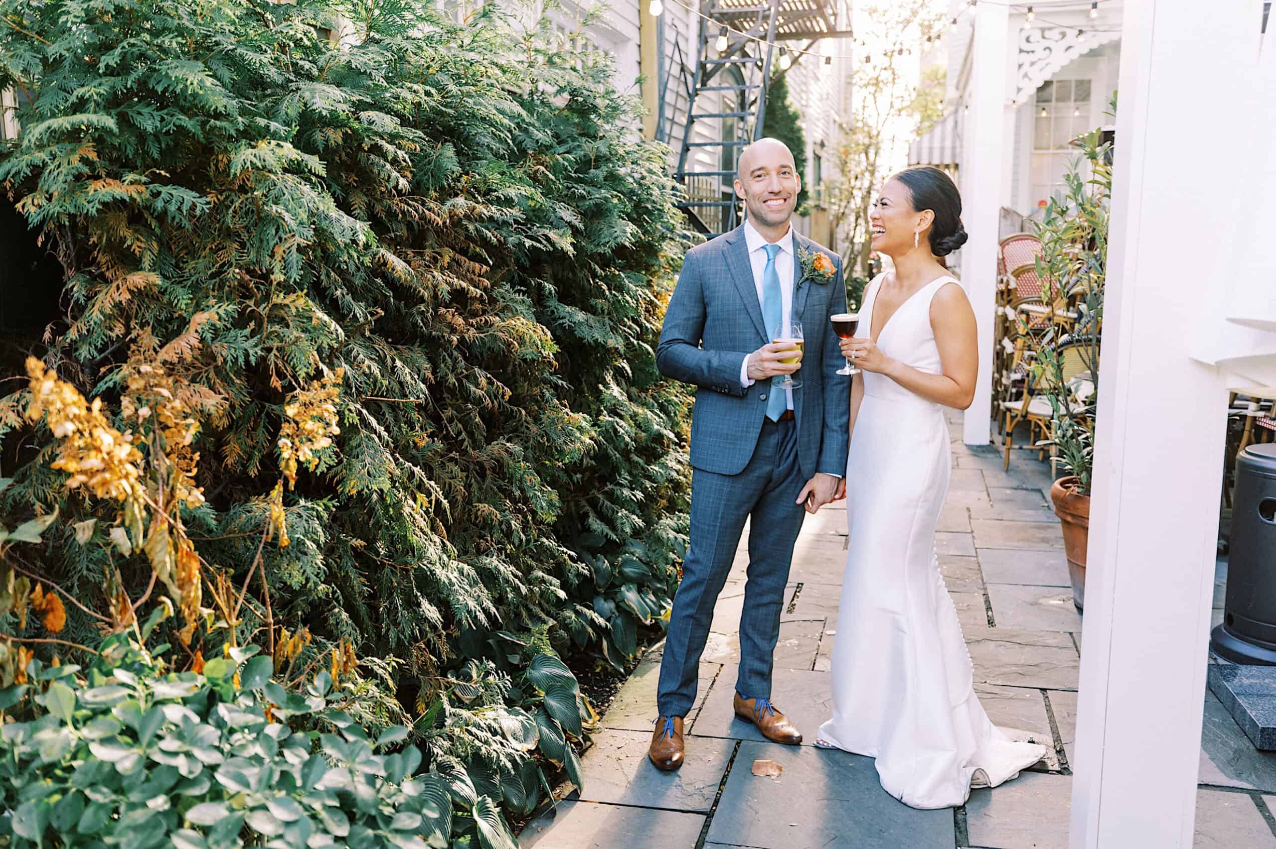 A man in a blue suit and a woman in a white dress stand outdoors on a stone walkway at a Cape May wedding at the Virginia Hotel, holding drinks and smiling at each other, surrounded by lush greenery.