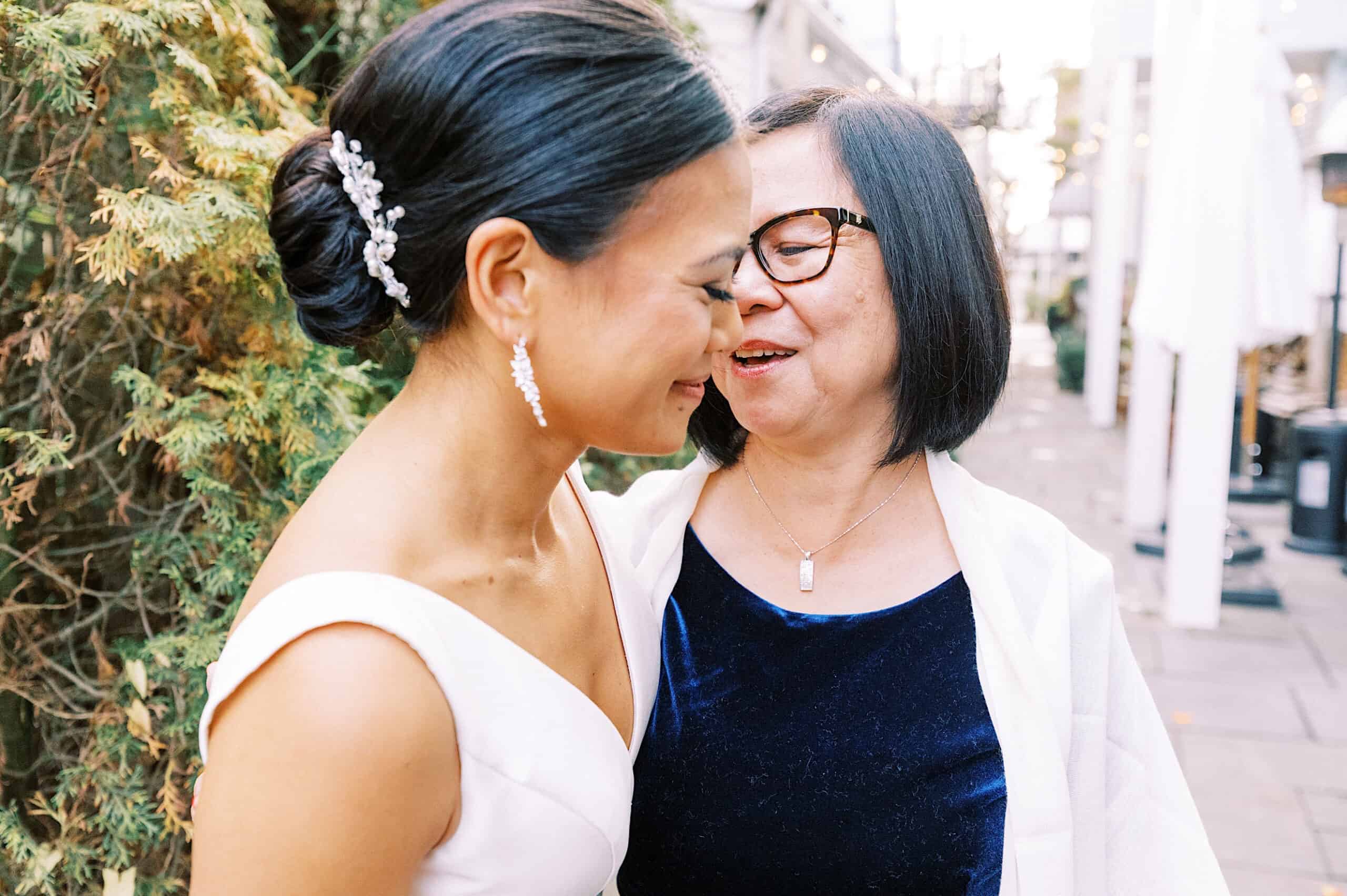 Two women, one in a wedding dress and the other in a blue dress with glasses, smile and stand close to each other outdoors during a Cape May wedding at the Virginia Hotel.