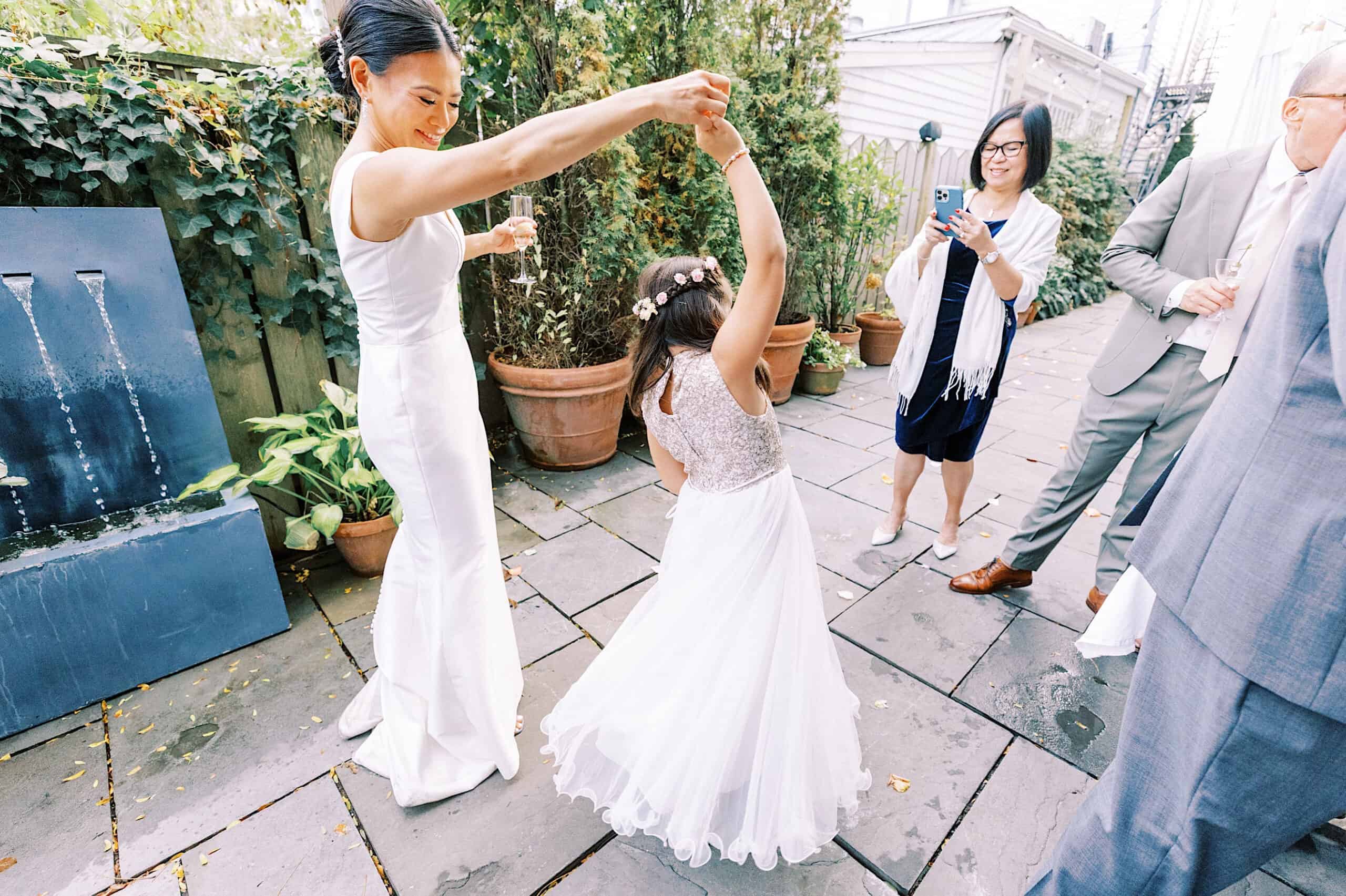 A woman in a white dress twirls a young girl on an outdoor stone patio during a Cape May wedding at the Virginia Hotel, while three adults watch and capture the joyful moment in photos.