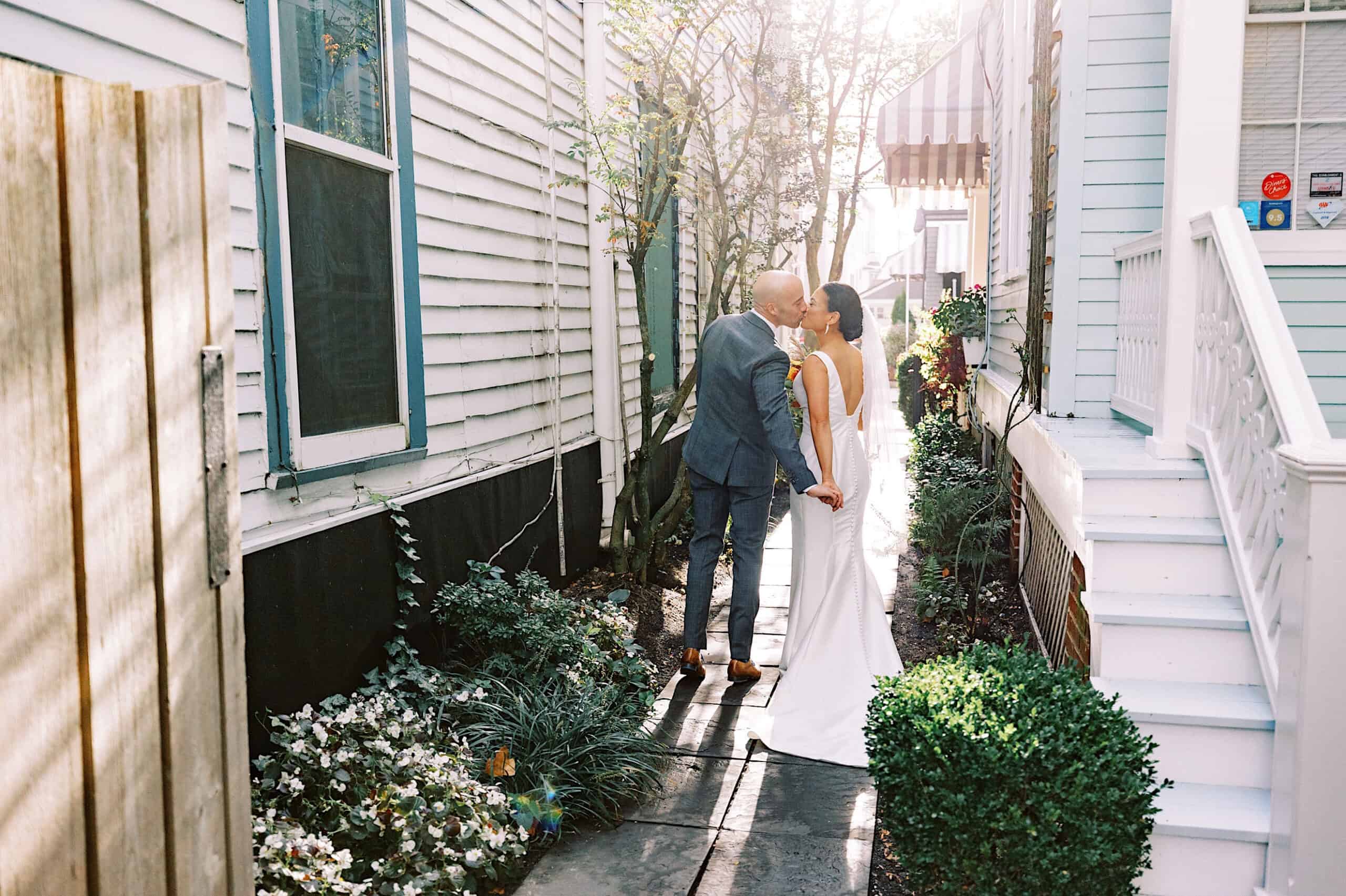 A couple in wedding attire holds hands and kisses while walking down a narrow outdoor path between two houses during their charming Cape May wedding at the Virginia Hotel.