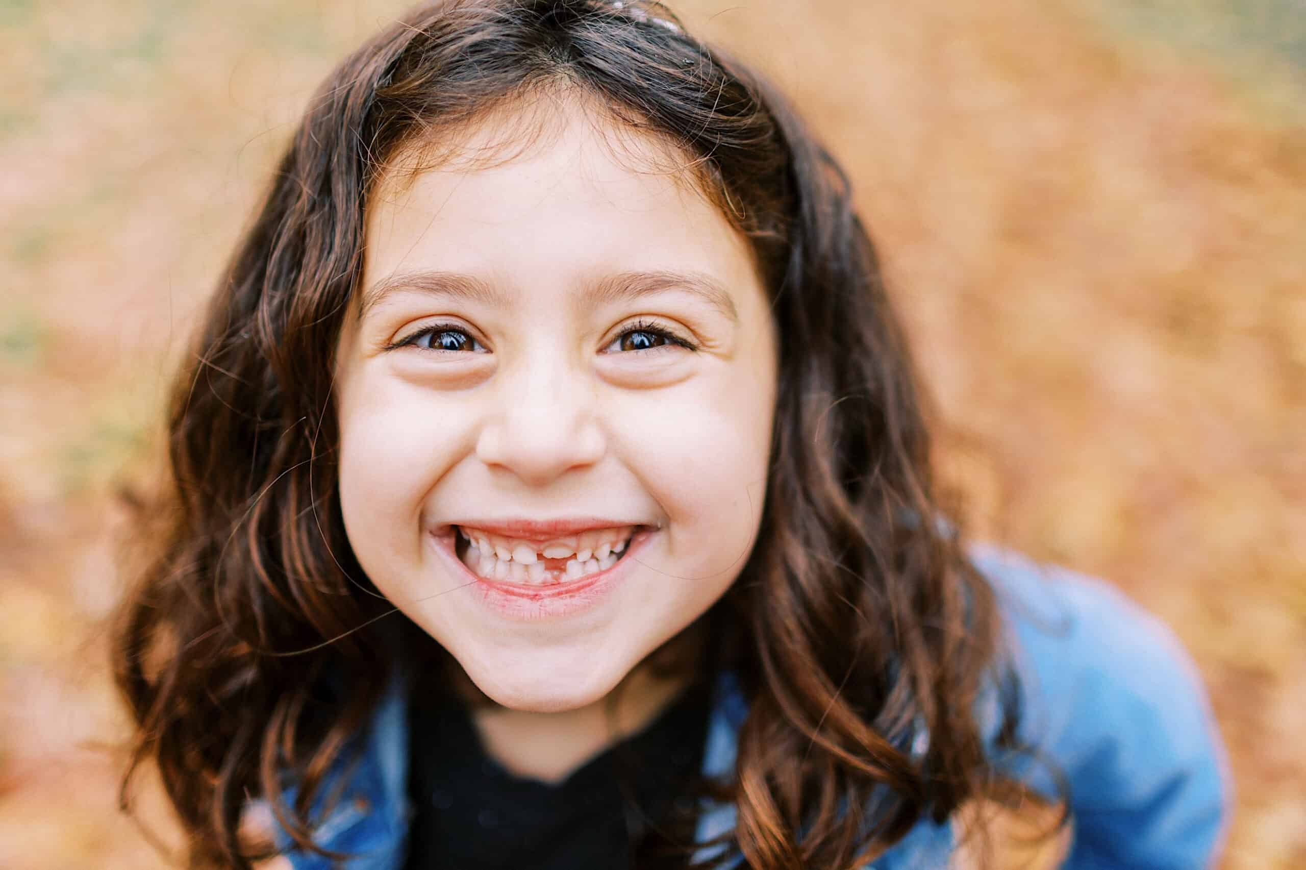 Young girl with brown hair smiles widely, showing missing teeth, while standing outdoors amid the autumn leaves during Fall Family Photos in Rittenhouse Square.
