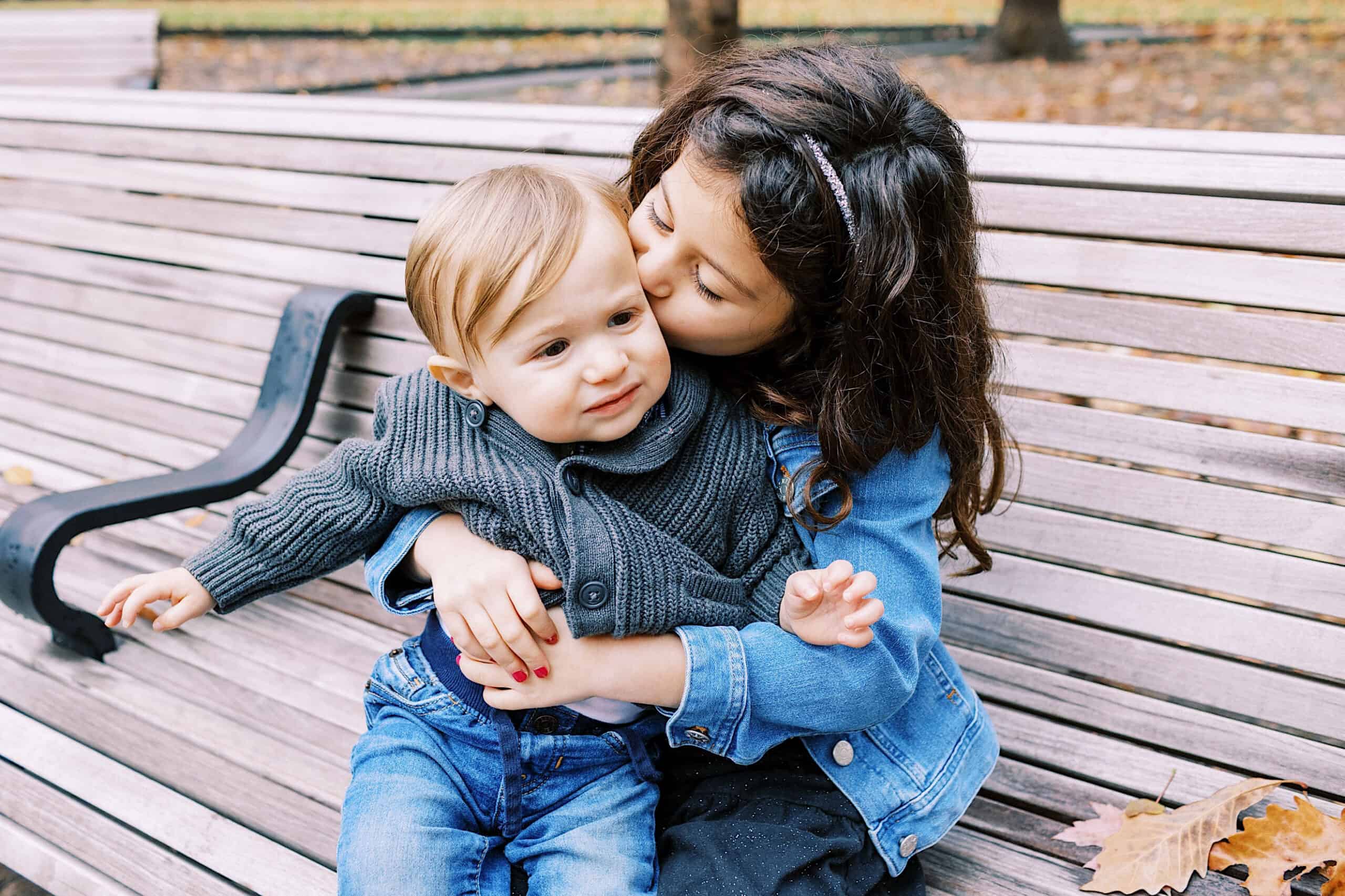 A young girl hugs and kisses a toddler on the cheek while sitting together on a wooden park bench in Rittenhouse Square; autumn leaves are scattered on the ground, capturing the warmth of fall family photos.