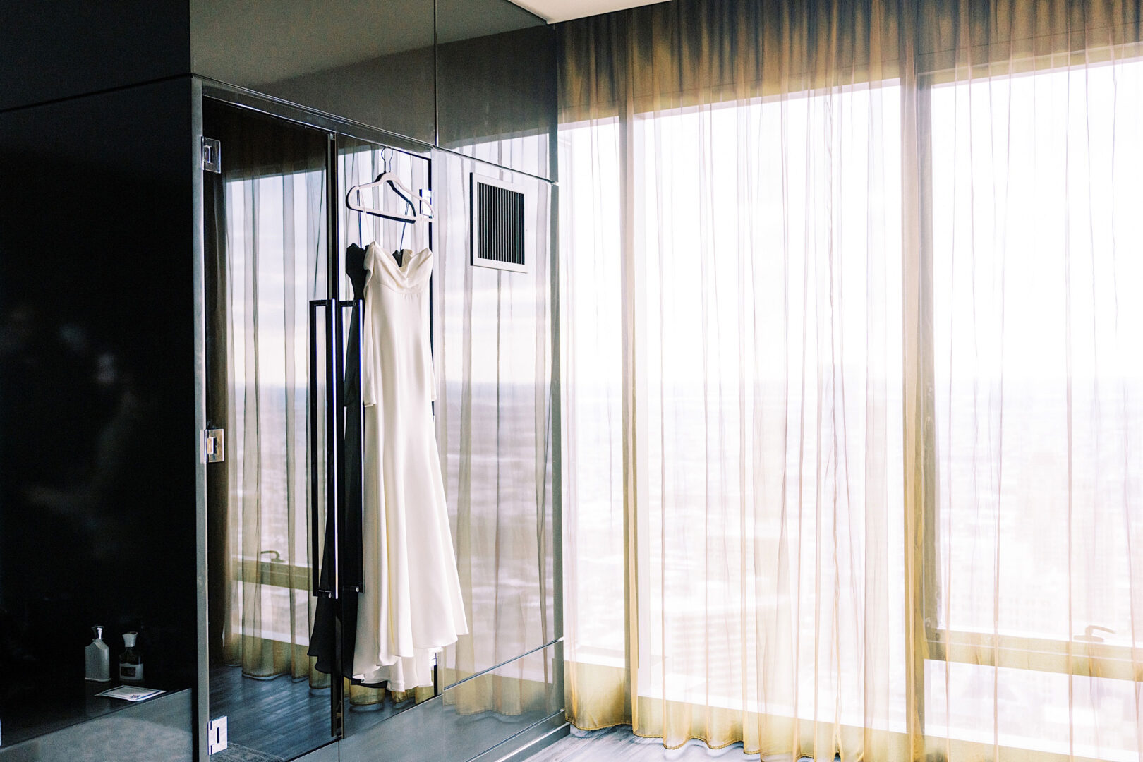 A white bridal gown hangs in a glass-enclosed shower area by a large window with sheer curtains, letting in soft natural light, as a gentle reminder of the New Year’s Eve wedding at the Kimmel Center in Philadelphia.