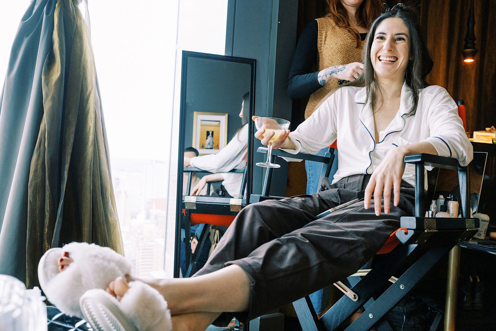 A woman in pajamas and slippers sits in a makeup chair, holding a drink and smiling, while a stylist with a tattooed arm works on her hair in preparation for New Year's Eve.