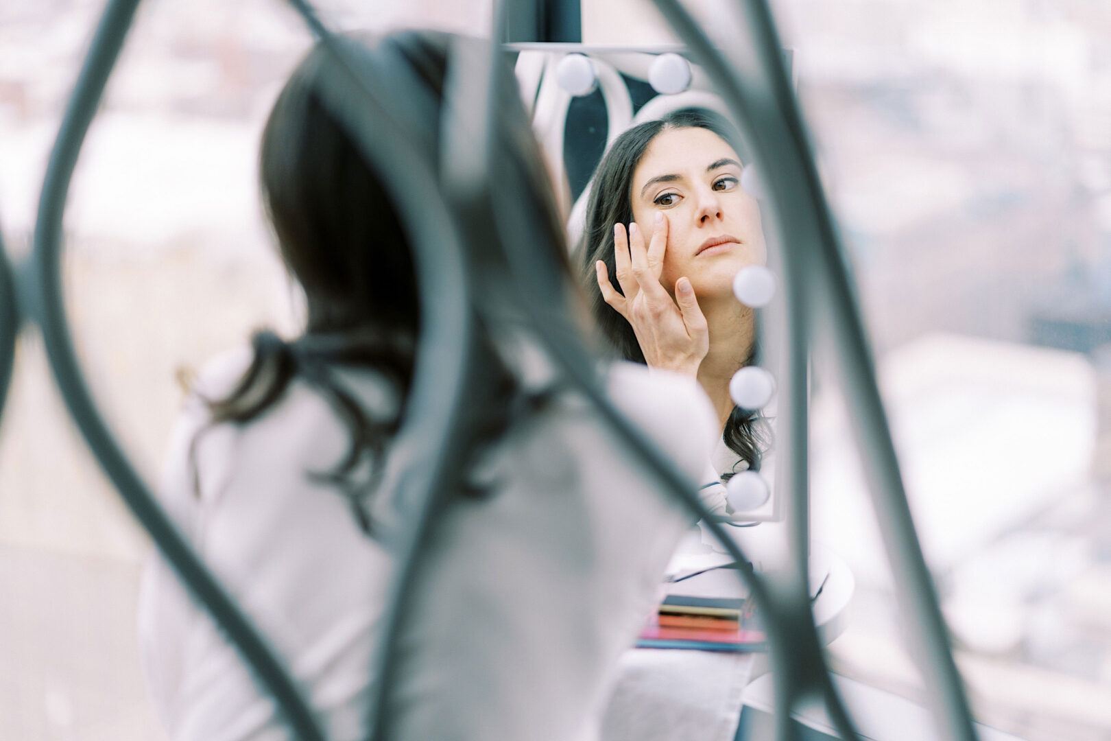 A woman applies makeup while looking at herself in a mirror with lights around it, preparing meticulously for a glamorous New Year's Eve celebration.
