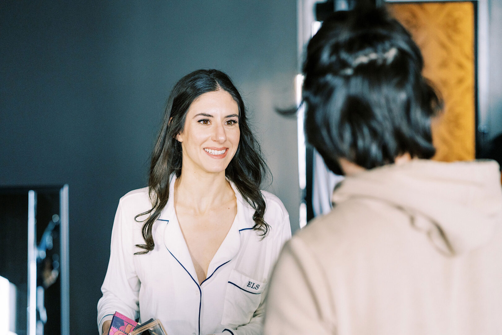 A woman with long hair, wearing a white shirt and holding a phone, smiles at another person in the foreground who is facing away from the camera at the Kimmel Center on New Year's Eve.