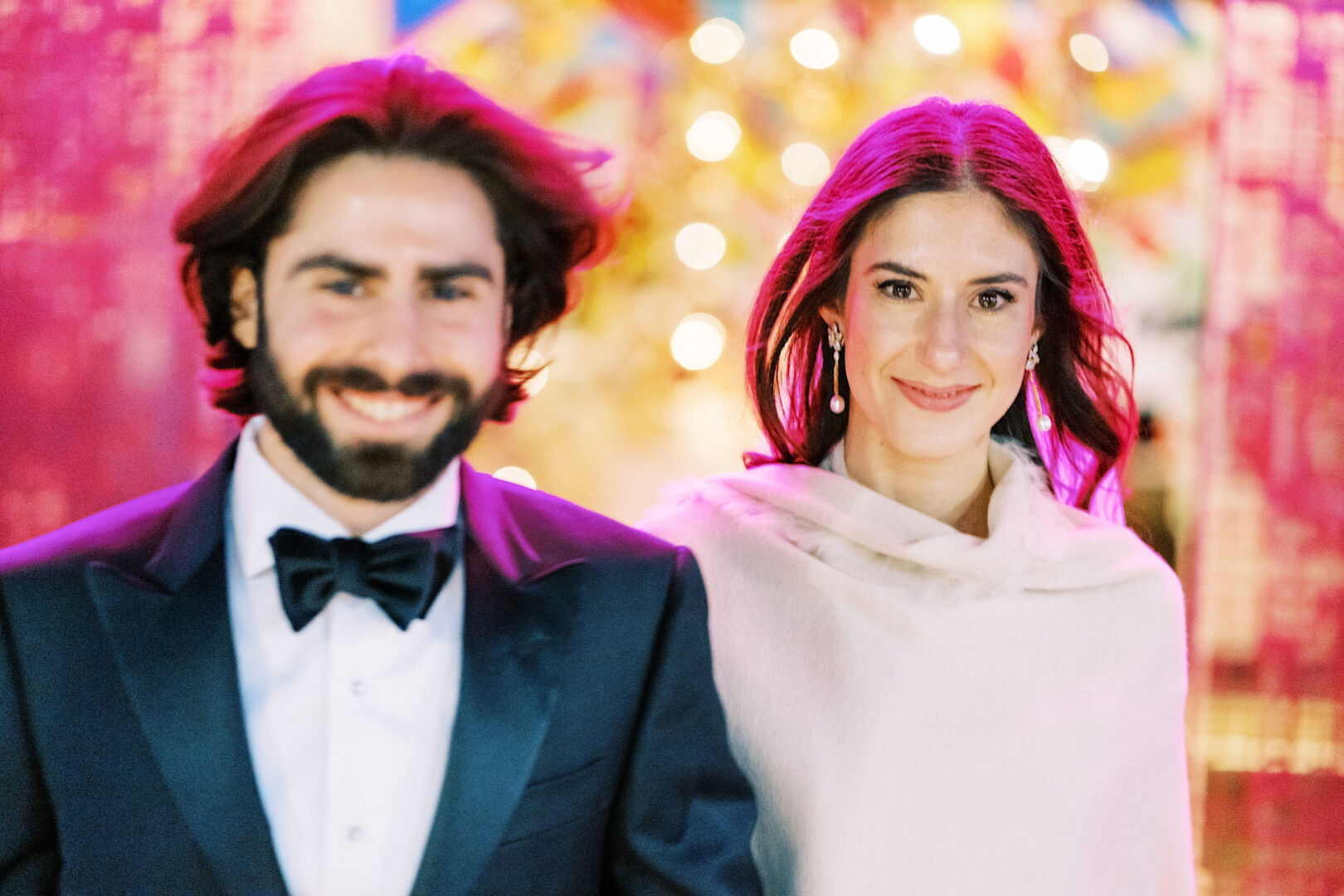A man in a black suit with a bow tie stands next to a woman in a light dress, both smiling at the camera in the brightly lit, colorful setting of the Kimmel Center during New Year's Eve celebrations.