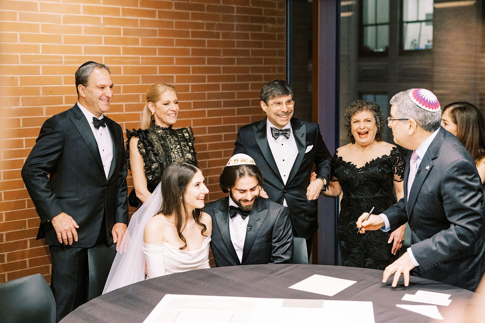 A bride and groom sit at a table with documents, surrounded by four standing individuals in formal attire and a person with a yarmulke, against a brick wall background. The festive setting hints at the Kimmel Center in Philadelphia on New Year's Eve.