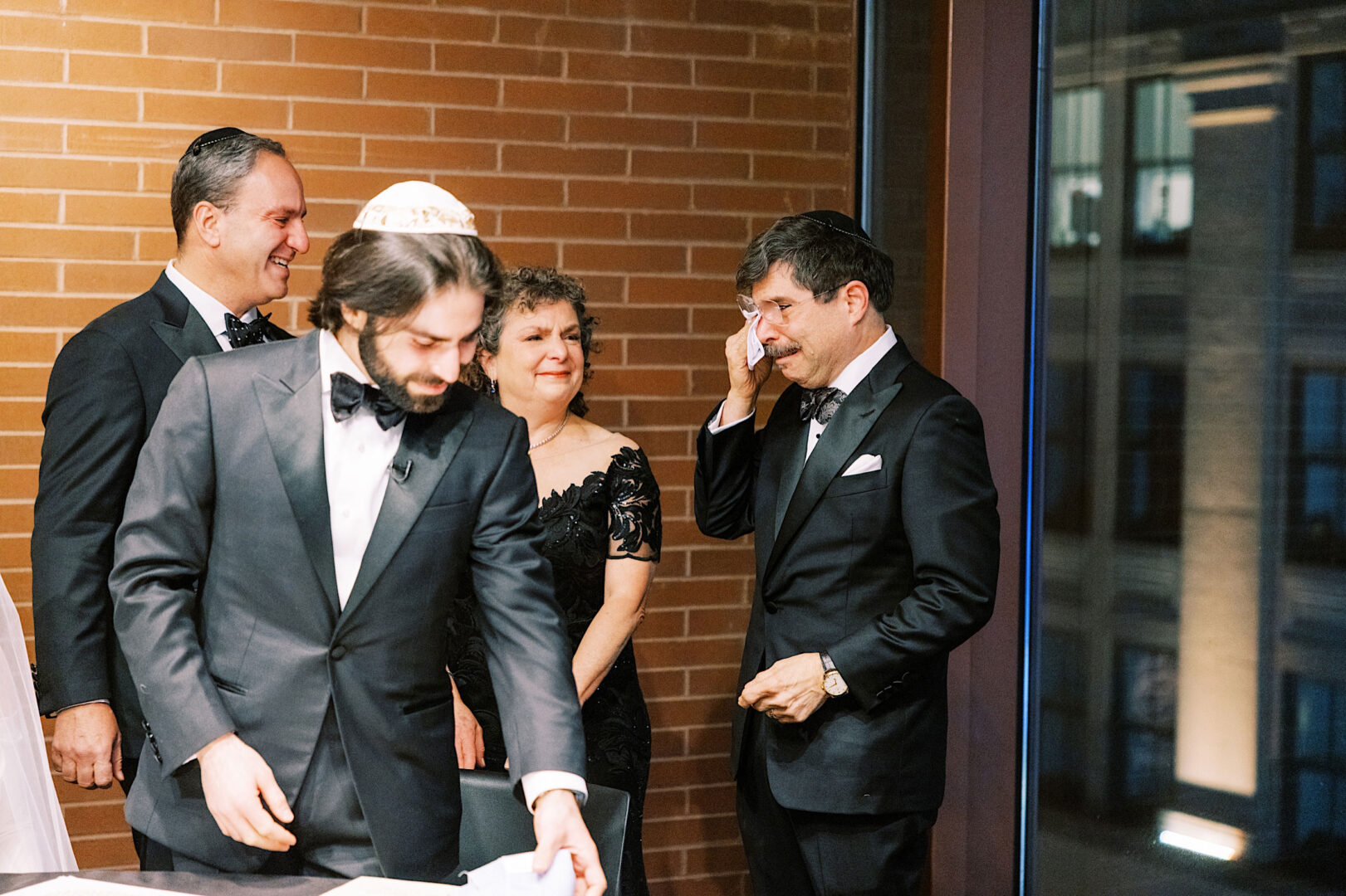 A group of formally dressed people stand together indoors in Philadelphia. Three men are in black suits and bow ties, and a woman in a black dress. One man appears emotional, holding a tissue to his eye, as if reflecting on a memorable New Year's Eve or perhaps a heartfelt wedding moment.